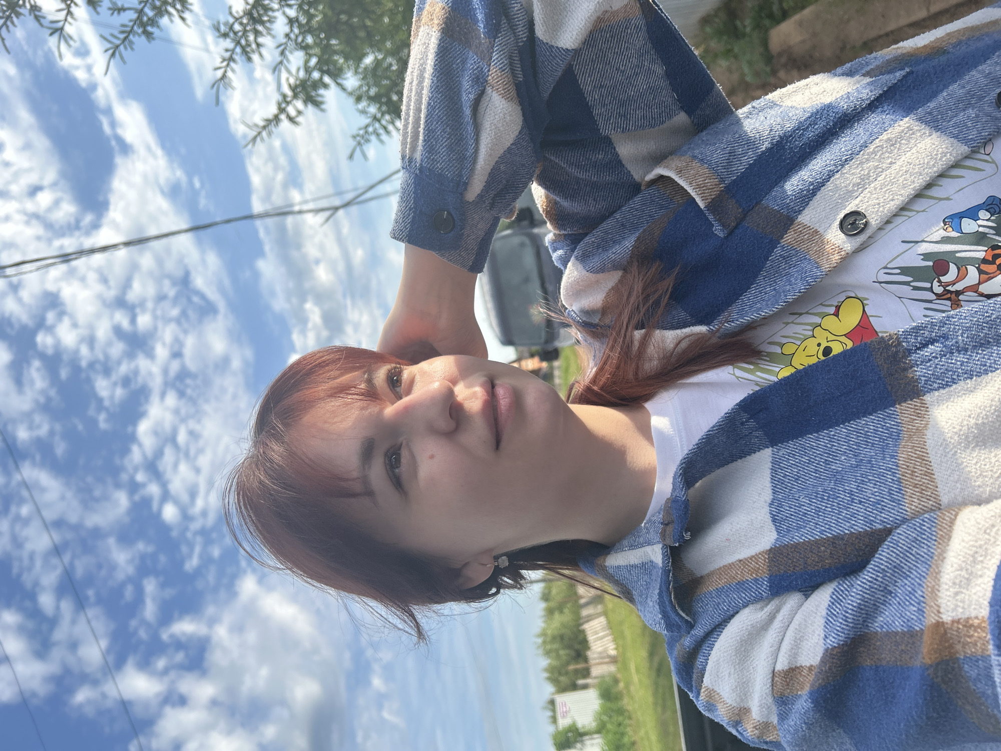 A young woman poses in an outdoor setting. She has reddish-brown hair and is wearing a white t-shirt with cartoon characters and a blue and white plaid flannel shirt. She is resting her head on her hand. The background features a bright blue sky with clouds. Green trees and other vegetation can be seen in the background. The location is most likely not Фоки, Russia, based on the scenery, which does not match the typical urban or industrial settings associated with that city. The background landscape suggests a more rural or suburban environment.