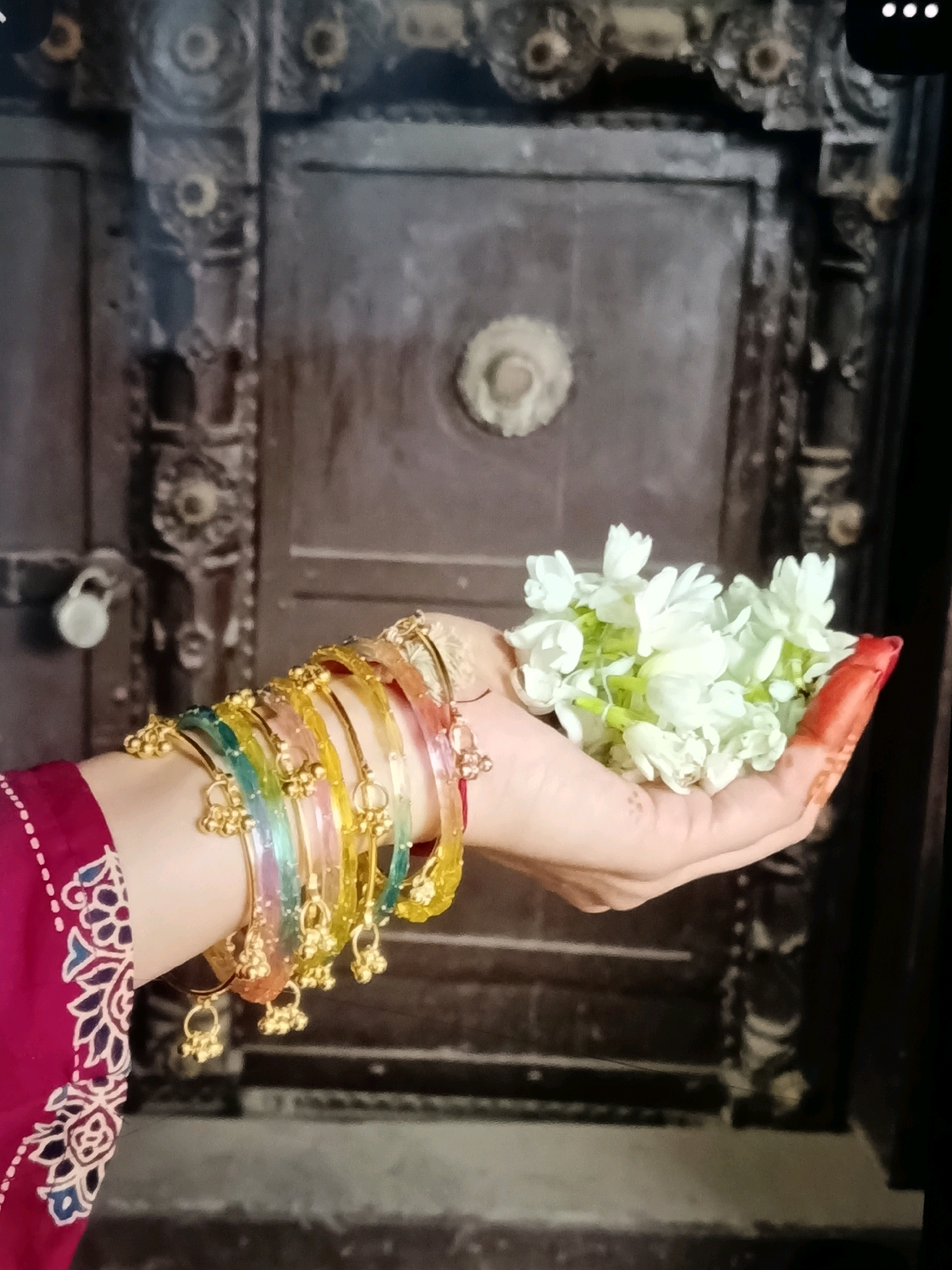 In C Camp, India, a close-up shot captures a hand adorned with colorful bangles and henna. The hand gently holds a cluster of delicate white flowers, possibly jasmine, against a backdrop of an antique wooden door. The door features intricate carvings and a prominent circular handle. A padlock is visible hanging on the left side of the door. The lighting is soft, casting subtle shadows and highlighting the textures of the bangles, flowers, and the aged wood.