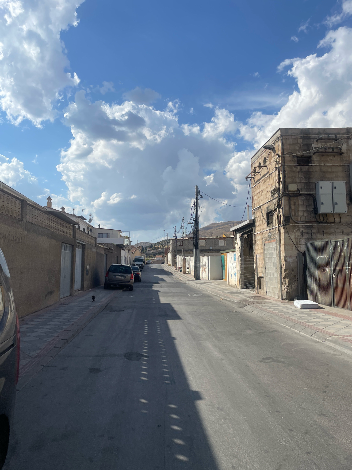 A street scene in Atarfe, Spain, shows a narrow asphalt road with a sidewalk on either side. The road is lined with low-rise buildings, some of which appear to be residential, with attached garages or storage areas. On the left side of the road, a dark-colored car is parked along the sidewalk, and a small black cat is walking on the checkered sidewalk. Further down the street, other vehicles are parked, including a white van. The buildings on the right side of the street are constructed from brick and stone, with visible electrical conduits and utility boxes attached to their facades. A metal gate is visible in front of one of the buildings. The sidewalk on the right side of the street has a concrete curb and a checkered tile pattern. A discarded mattress lies on the sidewalk near the right curb. In the background, rolling hills under a partly cloudy blue sky can be seen. The image is captured from a low angle, suggesting the photographer is standing on the street.