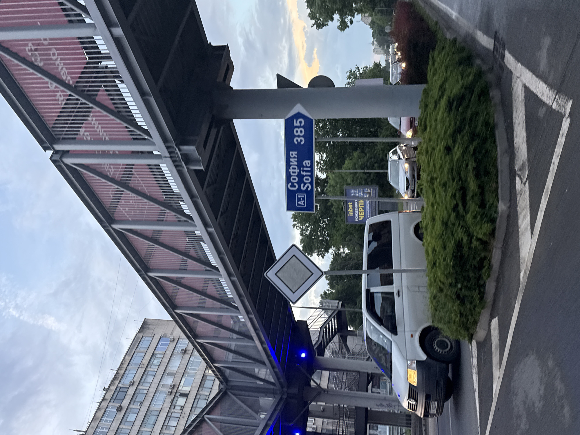The image shows a street scene in Burgas, Bulgaria. A pedestrian overpass spans the road, with blue accent lights illuminating its underside. A road sign indicates "София Sofia 385 A-1". Another sign with a white diamond is visible near the staircase of the overpass. A white van is parked near the sidewalk, adjacent to a hedge. In the background, there are other vehicles and a building. The sky is cloudy with a hint of sunset. The road has pedestrian crosswalk markings.