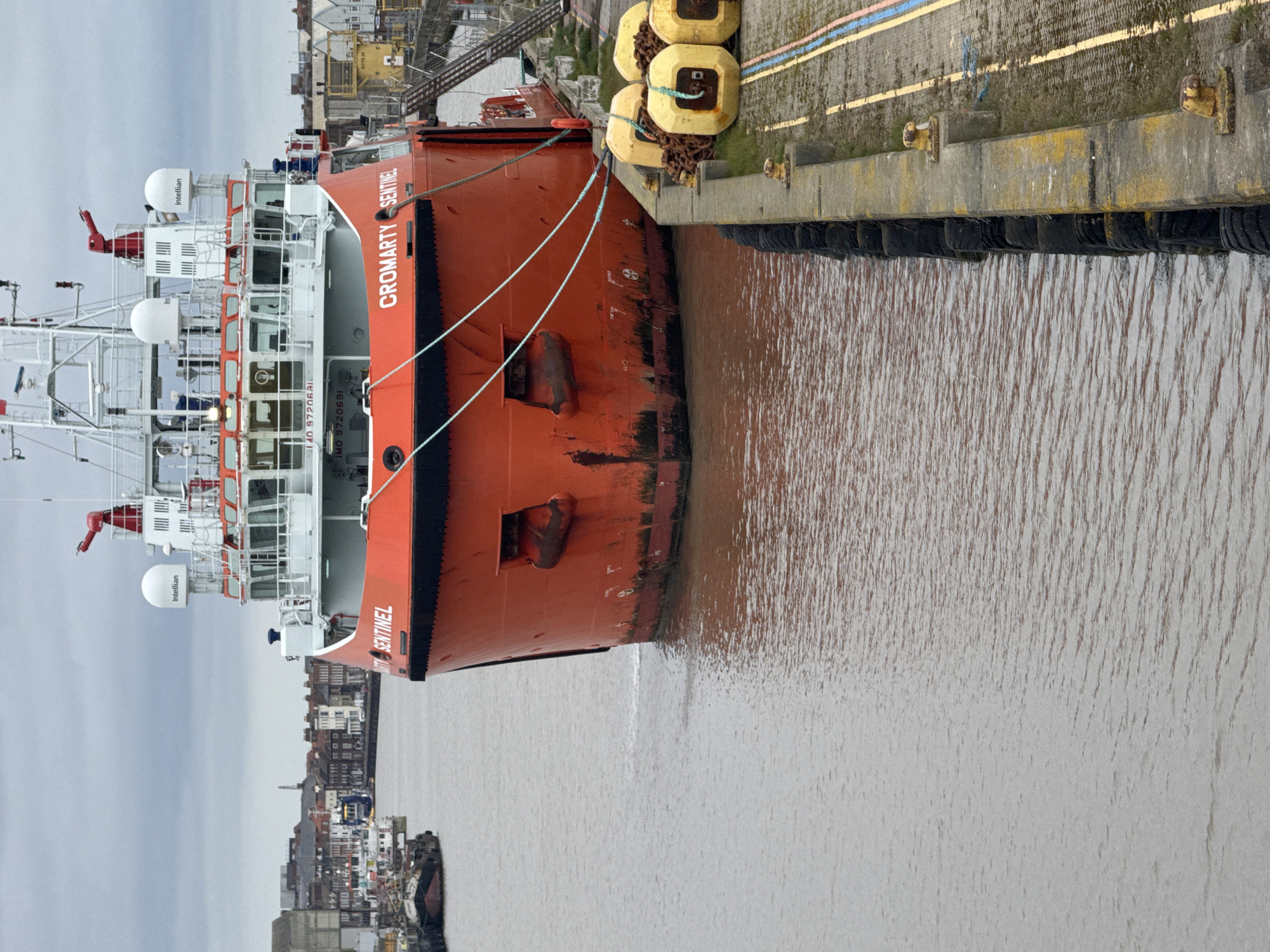 The image shows the port side of a large, red vessel named "Cromarty Sentinel" docked at a quay in Gorleston-on-Sea, United Kingdom. The ship is tied to the quay with mooring lines. The name "Sentinel" is visible near the bow. The ship's superstructure is white and contains navigation equipment. The quay is concrete and has black rubber tires hanging along its edge, and yellow buoys. The water is brown and muddy. In the background, the town of Gorleston-on-Sea is visible. Other industrial structures, including cranes and gantries, are visible in the background. The sky is overcast.