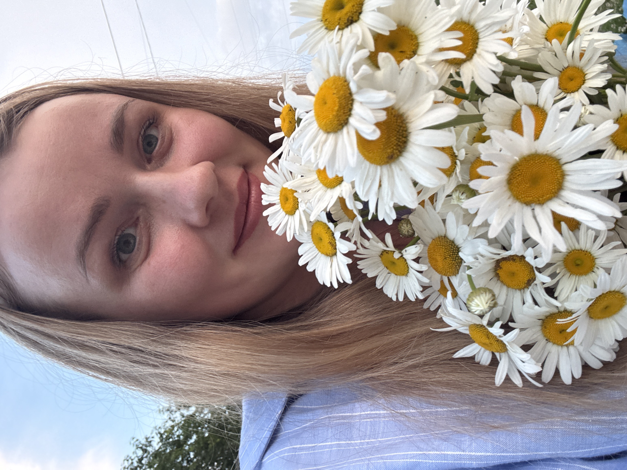 In this close-up, outdoor, selfie-style photograph, a young Caucasian woman with blonde hair is holding a bouquet of white daisies with yellow centers. She is wearing a light blue, pinstriped button-up shirt. Her face is turned slightly towards the camera, and she has a subtle smile. The background suggests a daytime setting with a cloudy sky and some greenery, possibly indicating a park or natural area. The image is likely taken in Chaikovsky, Russia, given the provided context.