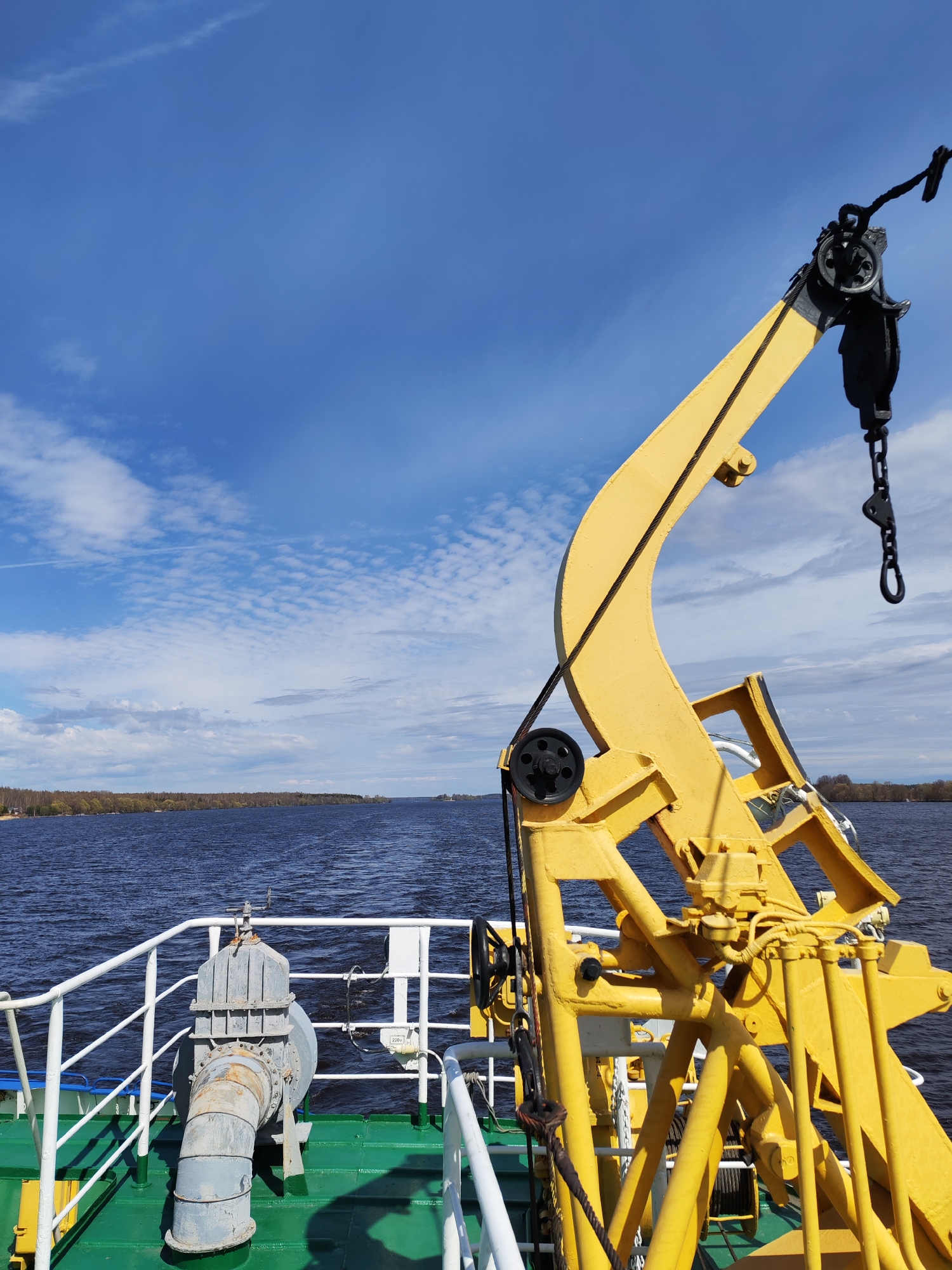 The image shows a view from the deck of a boat or ship, with a yellow crane dominating the right side of the frame. The crane has a pulley system and a chain hanging from its end. To the left, there's a gray industrial-looking piece of equipment with pipes and valves, secured to the green and white deck. A white railing runs along the edge of the deck.
The background features a body of water, possibly a river or lake, with a treelined shore in the distance. The sky is blue with scattered clouds. The location and city are not discernible from the image.