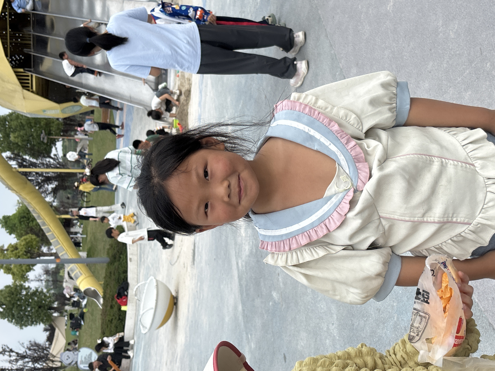 A young girl in a white and blue ruffled dress smiles at the camera while holding a bag of potato chips. The location appears to be a playground in Jiangjin, China, as evidenced by other people, playground equipment, and trees visible in the background. Children are playing on a yellow slide. Other adults and children are walking on the playground.