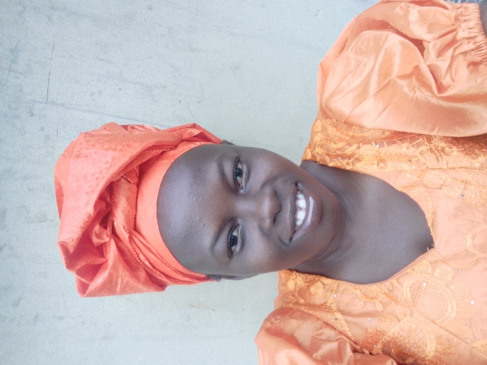 A woman with dark skin smiles warmly. She wears an orange head wrap and a matching blouse. The blouse features puffy sleeves and intricate lace detailing. The background appears to be a plain, light-colored wall. The lighting is bright and evenly distributed, highlighting her features and clothing.