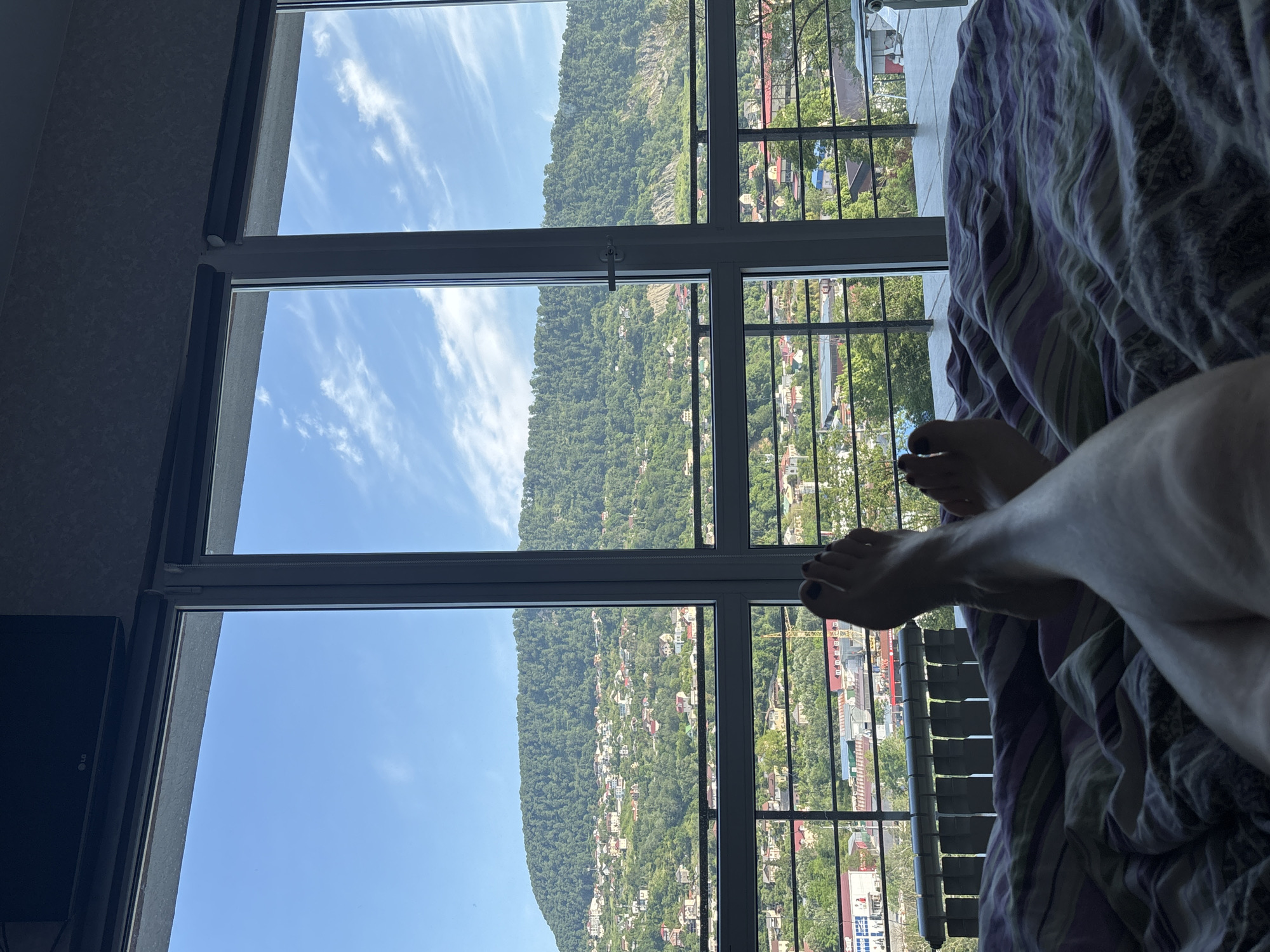 In this image, a person's bare legs and feet are visible, lying on a bed with striped bedding. The perspective is from the bed, looking out through a large window. The window offers a view of a lush green mountainside dotted with houses, characteristic of the Khostinsky District of Sochi, Russia. Below the mountain, a cityscape with buildings and trees is visible, separated by a balcony railing. The sky is blue with scattered white clouds. An air conditioning unit is mounted on the wall above the window.