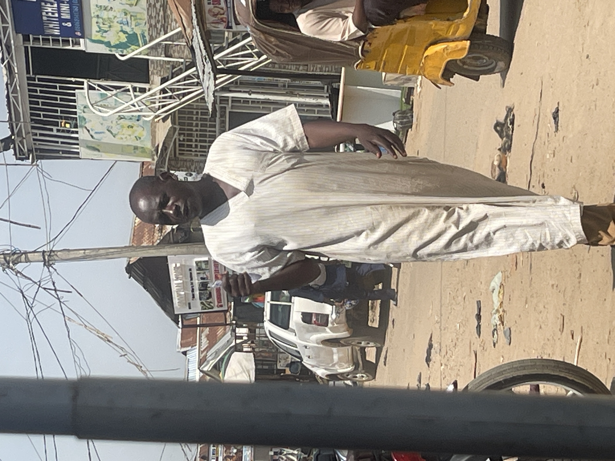 The image depicts a street scene in Abuja, Nigeria, on a sunny day. A man in a light-colored, striped robe walks toward the right, holding a small object in his right hand. Behind him, a white pickup truck is parked on the dusty street. Further down the street, a yellow motorized tricycle, commonly known as a "Keke Napep," is visible with a person sitting inside. Buildings with corrugated iron roofs and signs are present in the background, indicating a commercial or residential area. The ground is unpaved and scattered with debris. Overhead, electrical wires crisscross against the bright sky.