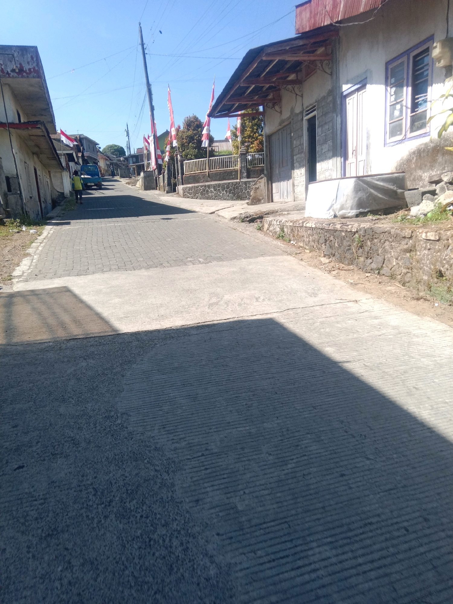 The image shows a street in Kapencar, Indonesia, on a sunny day. The street is paved with cobblestones in the background and a smoother concrete surface in the foreground. Buildings line the street on both sides. On the right, a house with a white facade, a red corrugated metal roof, and purple-trimmed windows is visible. A stone wall with some vegetation runs along the front of this property. On the left, another building with a white facade and a weathered appearance is present.
Several Indonesian flags, red and white, are displayed on poles along the street, suggesting a patriotic occasion. A small blue truck is parked on the left side of the street, and a person in a yellow shirt is standing near it. The sky is clear and blue. The scene appears to be a typical residential street, possibly in a rural or semi-urban area. The overall impression is one of a quiet, ordinary day with a touch of national pride.