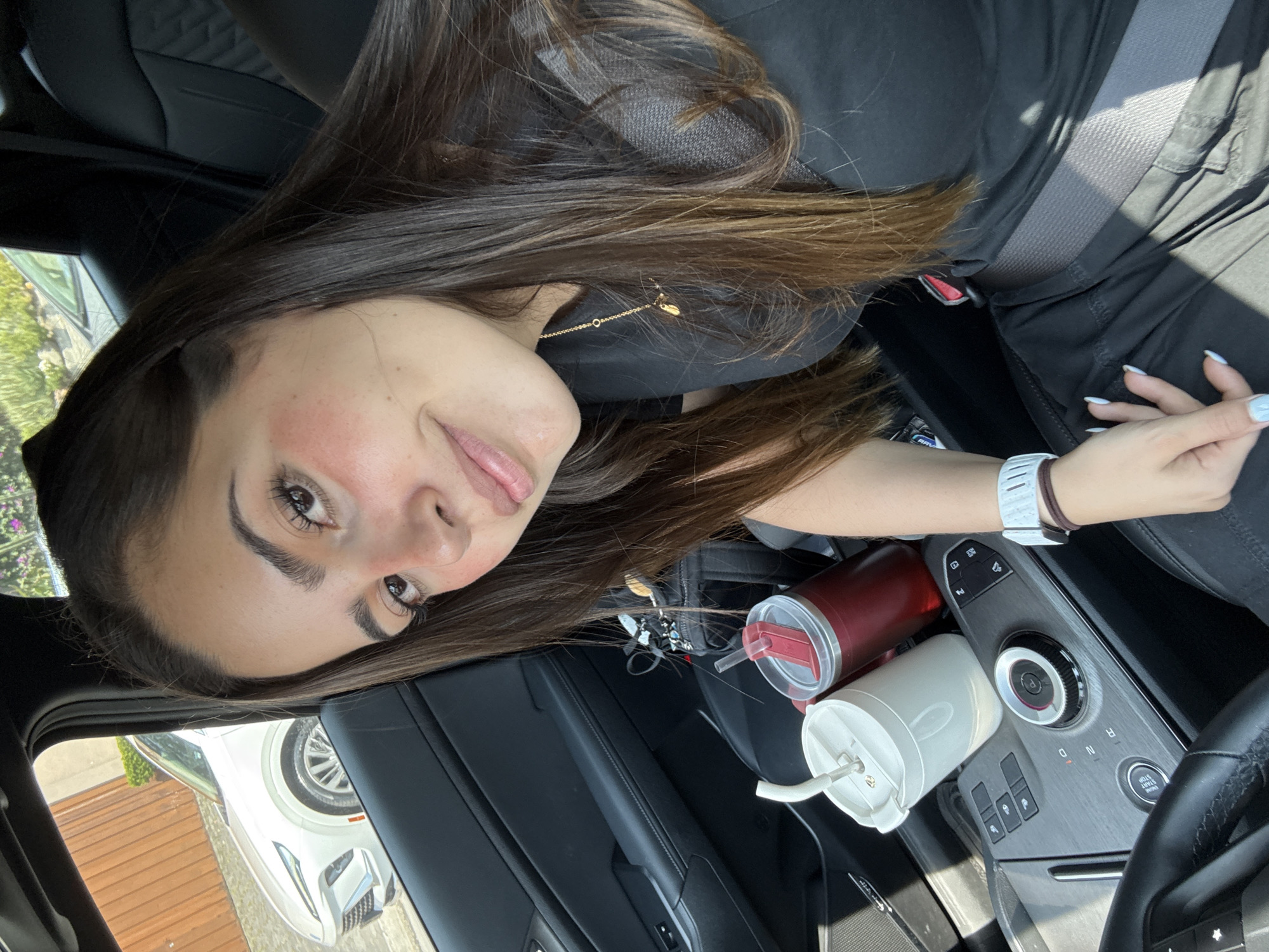 A woman with long brown hair and a light complexion is in the driver's seat of a car. She is wearing a black top and a seatbelt. Her left hand is visible, resting near the center console. Two cups are in the cupholders. The interior of the car is black. A white vehicle is visible through the car window. The location is not discernible as Puebla, Mexico from the content of the image alone.