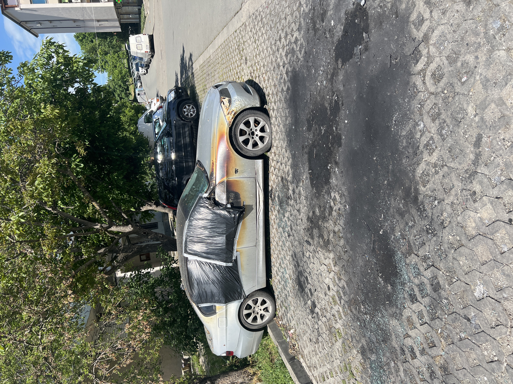 A silver sedan car, seemingly burned out, is parked on a cobblestone street. The vehicle's windows are covered with black plastic sheeting. Burn marks are visible on the hood and driver's side. Shards of glass are scattered on the pavement next to the car. A black SUV is parked directly behind the silver car. Further down the street, several white vans are also parked. Trees line the side of the street, with a building visible in the background. Based on the environment, the location is likely in Burgas, Bulgaria. There are no people visible in the frame.