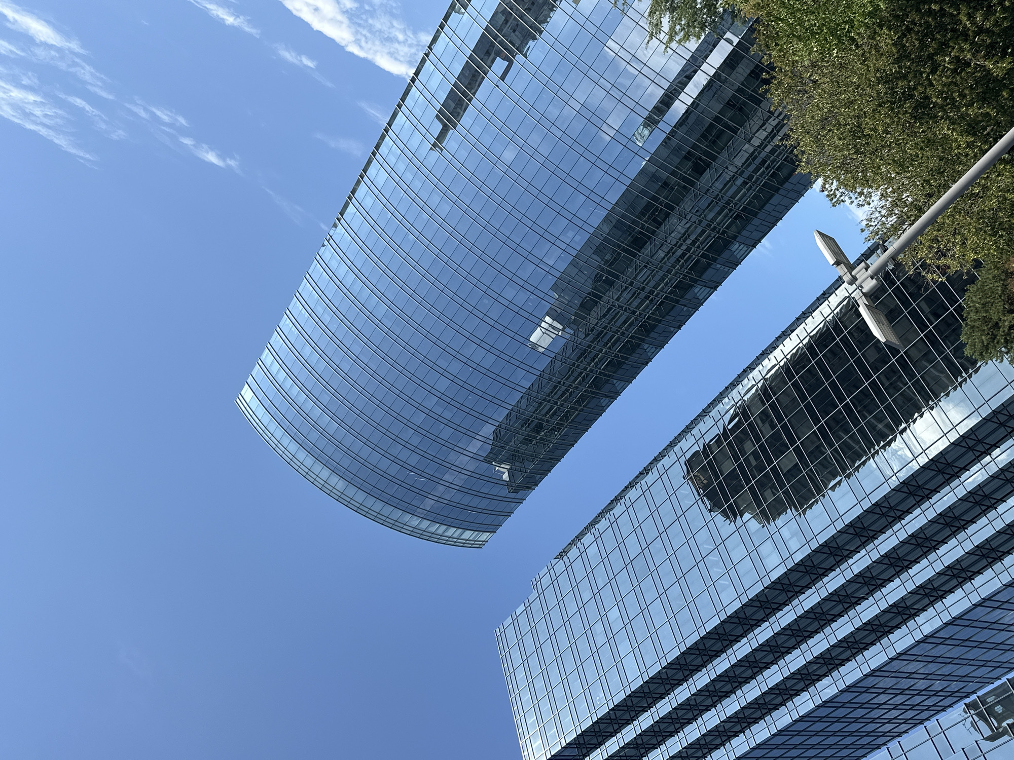 This is a low-angle, close-up shot of two modern, glass-facade skyscrapers against a clear blue sky with scattered wispy clouds. The buildings are tall and sleek, with reflections of other buildings and the sky visible on their surfaces. The left building is rectangular, while the right building has a curved, cylindrical design. In the lower right portion of the image, a street lamp on a grey pole extends upwards, and lush green trees frame the bottom edge of the skyscrapers. The scene suggests a bright, sunny day in an urban environment. This image appears to have been taken in the Metrotown area of Burnaby, Canada, given the architectural style and the presence of prominent glass towers often associated with that district. There are no people or visible actions depicted in the image.