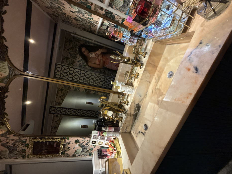This image depicts a luxurious bathroom interior at the Faena Hotel Miami Beach, United States of America. The scene is dominated by a long, light-colored marble trough sink with two drain grates. Above the sink are two arched mirrors with ornate gold frames. The walls are covered in pink wallpaper featuring a repeating pattern of zebras and tropical foliage.
In the reflection of the right mirror, a female subject with long, dark hair is visible, holding a smartphone and appearing to take a mirror selfie. She wears a light-colored, patterned top.
On the marble countertop, multiple ornate gold-colored fixtures are present, including four faucets resembling stylized animal heads and two decorative soap dispensers/holders. To the left of the sink, there is a stack of white items, various cosmetic products, a golden box labeled "LIPS," and a small golden tray holding several red-topped lollipop-like items. To the right, a multi-tiered gold metal rack displays numerous perfume bottles of varying shapes and colors, alongside a small glass container with dried botanicals. The lower portion of the image reveals dark-colored cabinetry beneath the sink. The room is brightly lit, with reflections visible on polished surfaces.