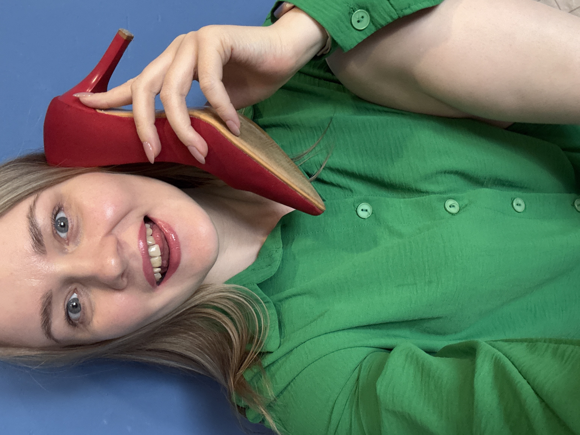 The image depicts a woman lying on a blue surface, presumably indoors. She is wearing a green button-down shirt and holding a red high-heeled shoe near her head. The woman is smiling and looking directly at the camera. The photograph does not offer any clues pertaining to the city of Chaikovsky, Russia.