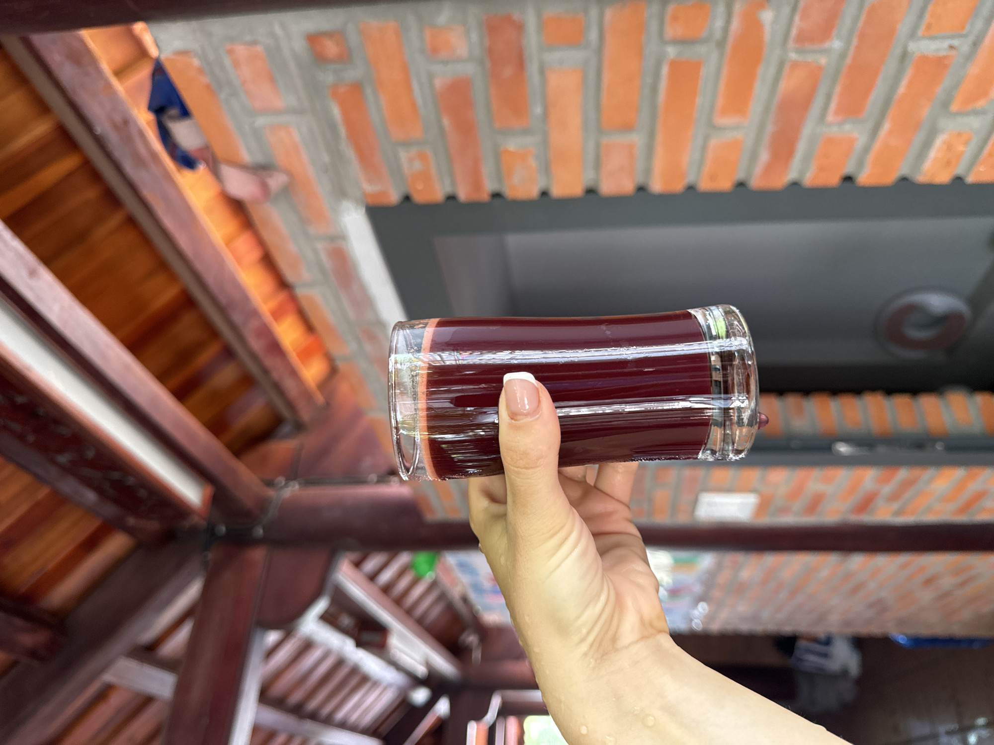 In Mũi Né, Vietnam, a hand with manicured nails holds a glass of dark red juice. The glass is cylindrical and appears to be made of clear glass, with a ridged texture on the exterior. The juice fills most of the glass and has a deep, opaque color. The hand is lightly moist, with small water droplets visible on the skin.
The background suggests an outdoor or semi-outdoor setting. Above, a wooden roof structure with dark brown beams and orange-tiled roofing is visible. To the right, a brick wall composed of orange-colored bricks and gray mortar forms a significant portion of the background. Behind the brick wall, a dark gray surface with a circular vent or decorative element is partially visible. The overall lighting suggests it is daytime.