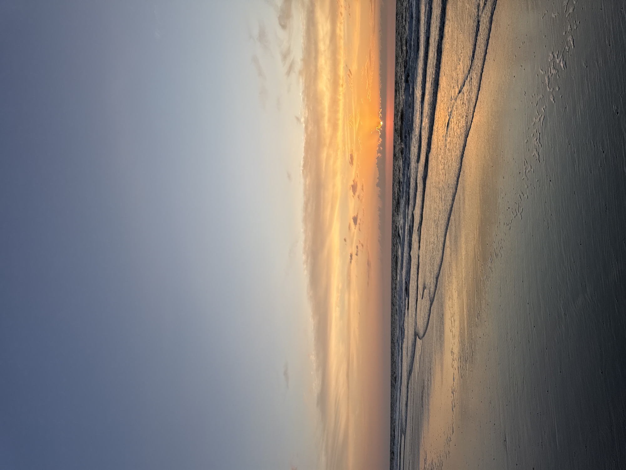 The image depicts a serene beach scene at sunset on the island of Langeoog, Germany. The sky displays a gradient of colors, transitioning from a pale blue at the top to vibrant oranges and yellows near the horizon. Wispy clouds are scattered across the sky, catching the warm hues of the setting sun. The sun itself is partially obscured by clouds, peeking through with its radiant rays extending outwards. Below the sky, the ocean meets the shore with gentle waves creating a foamy white line as they recede. The wet sand reflects the colorful sky, creating a mirror-like effect. The beach appears smooth and vast, with subtle textures from the sand and ripples left by the receding tide. There are no people or structures visible in the scene, emphasizing the natural beauty of the coastal environment. The overall impression is one of tranquility and natural splendor, capturing the quiet beauty of a coastal sunset.