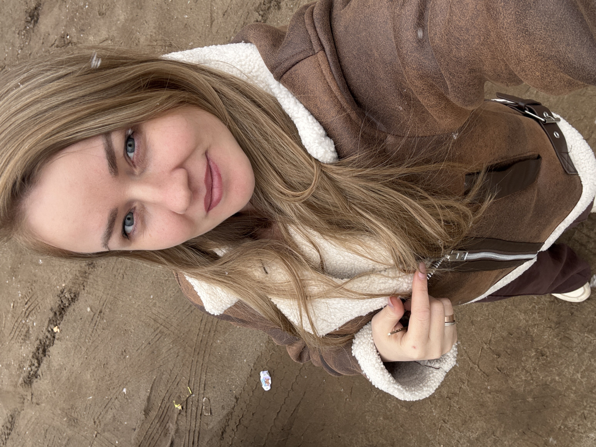 A young woman is standing on a sandy or earthen surface, possibly a beach or construction area. She is fair-skinned with blonde hair, blue eyes, and is wearing a brown suede jacket with white fleece lining. She is holding the jacket zipper with her left hand, which has two rings on the ring finger.
The ground is covered with tire tracks and small pieces of debris. Based on the tire tracks and the sandy ground, the location could be near a construction site or an unpaved area near a beach.
Given the sandy/earthen surface and lack of distinct landmarks, precise location identification in the city of Chaikovsky, Russia, is not possible without further information.