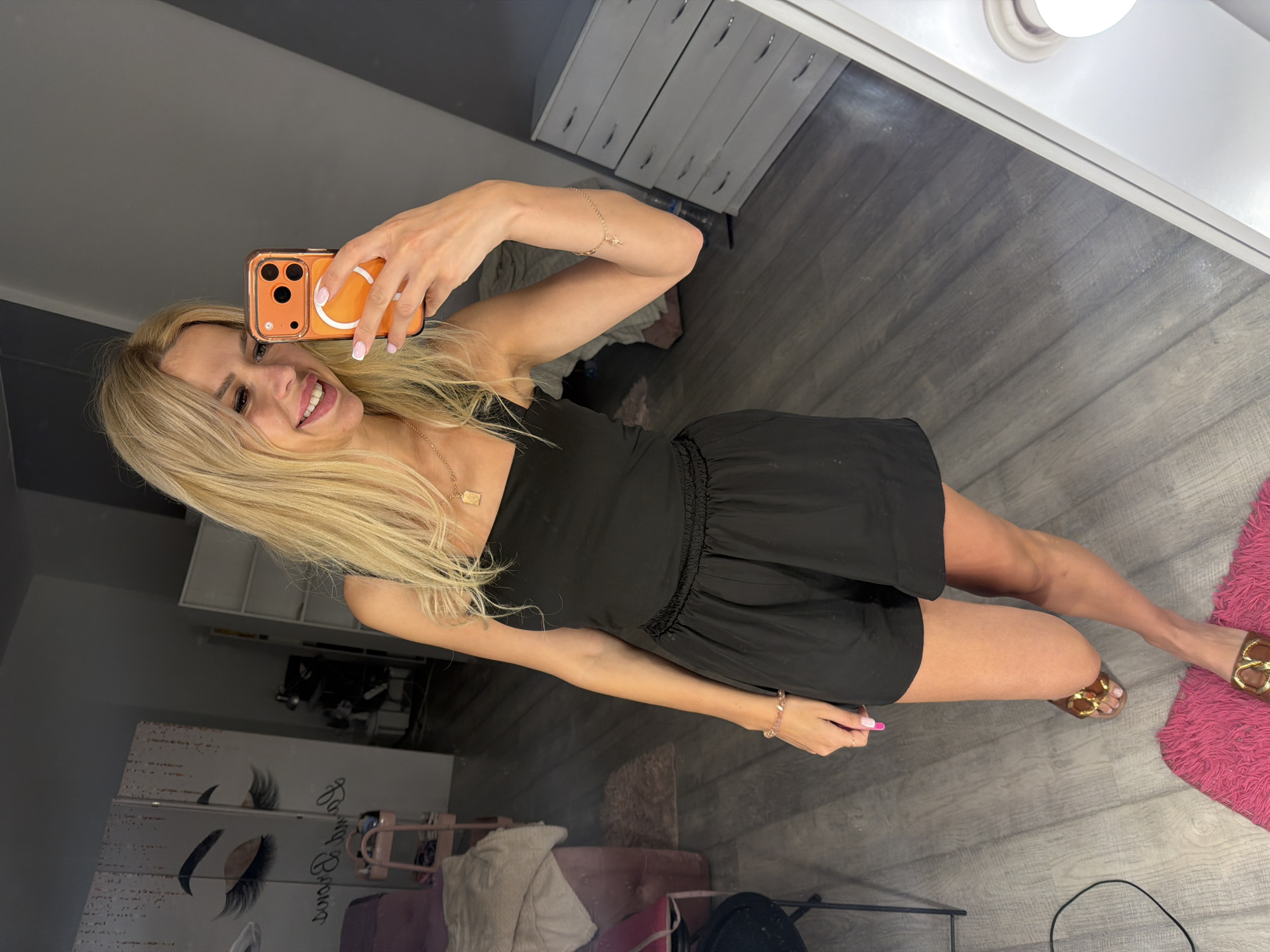 A woman with blonde hair is taking a mirror selfie in what appears to be a room with grey wooden flooring and a white mirrored closet in the background. She is wearing a black dress and brown sandals, and is holding a smartphone with an orange case. The room has feminine decor, including a pink rug and what looks like a makeup station. The woman is smiling and appears to be in good spirits. The image is taken from a slightly high angle.