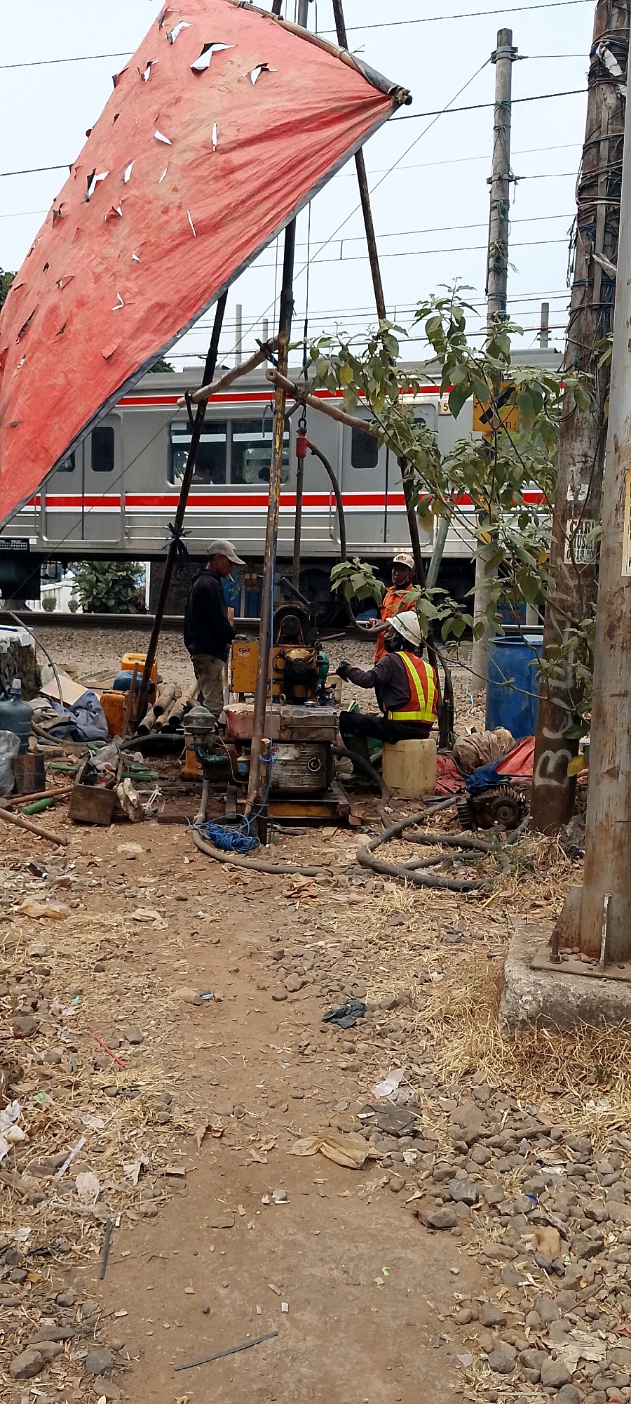 The image depicts an outdoor scene, likely in Bekasi, Indonesia, near a railway track. A damaged red tarpaulin is stretched over a wooden frame, providing shade. In the foreground, there is a construction or drilling site with various tools and equipment scattered on the ground, including pipes, hoses, a generator, and large blue barrels. Three individuals are present. One man, wearing a light-colored cap and dark clothing, stands near the equipment, smoking a cigarette. Another man, in an orange uniform and a white hard hat, is operating a machine. A third person, also in an orange uniform and white hard hat with a reflective vest, is seated and appears to be working on the equipment. A train with red and silver stripes is visible on the railway tracks in the background, along with utility poles and overhead wires. The ground is uneven and covered with dirt, gravel, and debris, including plastic waste. The overall impression is one of ongoing work in a somewhat unkempt, industrial or semi-industrial environment.