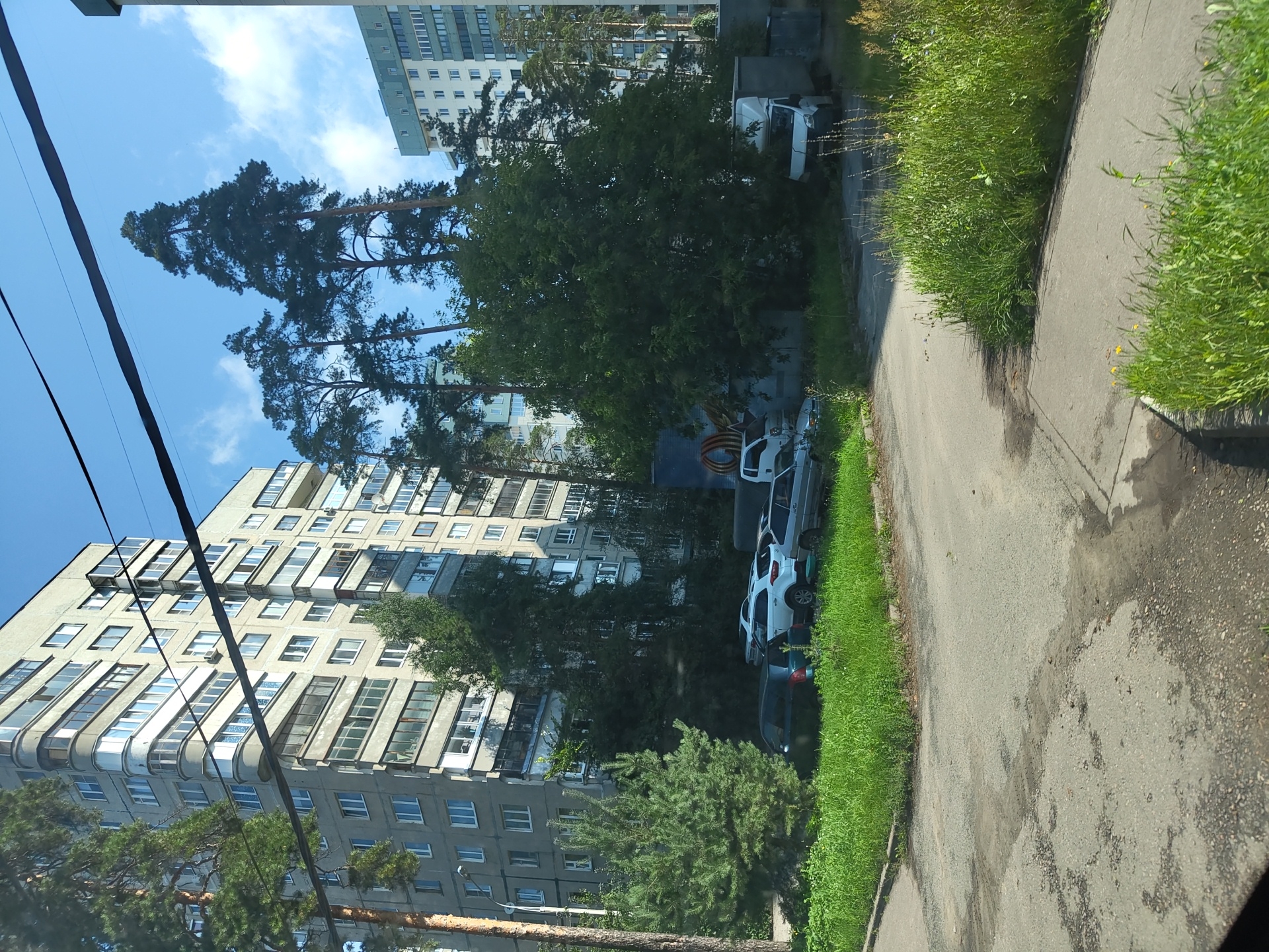 A daytime, eye-level, outdoor shot captures a street scene in Zhukovsky, Russia, featuring residential buildings, parked vehicles, and vegetation. Tall, slender pine trees and deciduous trees are prominent, interspersed with apartment buildings. Several cars, including a white SUV and a silver sedan, are parked on a paved area adjacent to a grassy strip. A white cargo van is also visible. The sky is blue with scattered white clouds. The foreground shows a worn asphalt road with some puddles. The image appears to be taken from a slightly elevated perspective, possibly from a vehicle.