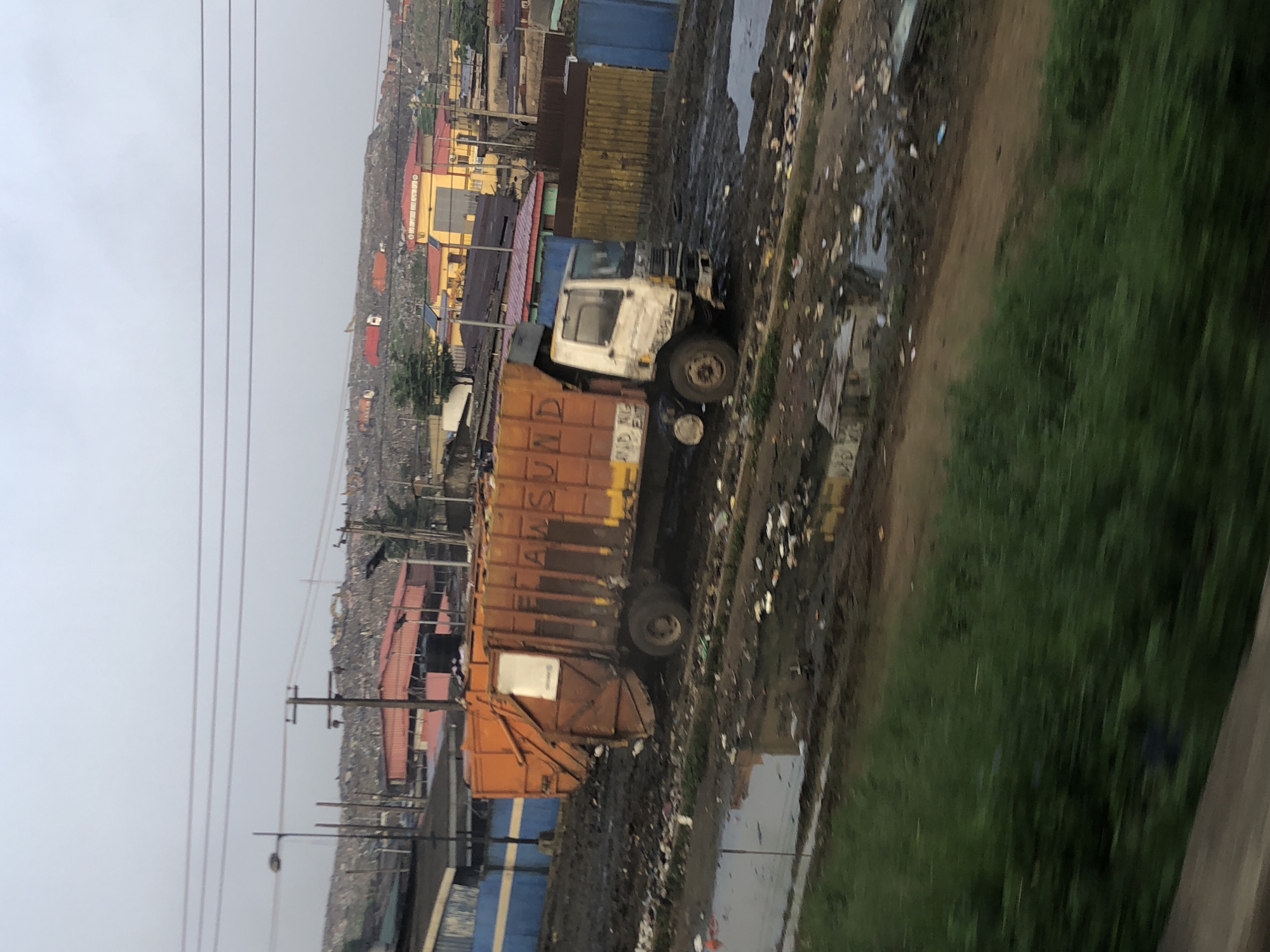 This is a medium-range, eye-level, outdoor shot taken on an overcast day in a densely populated, informal urban settlement, likely a landfill or recycling area, in Ikeja, Nigeria. The foreground is dominated by a muddy, debris-strewn path, with patches of stagnant water reflecting the dull sky. Green vegetation, blurred due to motion, occupies the bottom left of the frame.
In the mid-ground, an orange and white garbage truck is positioned at an angle, its rear loader extended and appearing to be in operation. The side of the truck has lettering that is partially visible, reading "LAW SUND." The ground around the truck is heavily muddied and littered with various refuse. Behind the truck, a series of low-rise buildings with colorful roofs (predominantly red and yellow) are visible, suggesting a mixed residential and commercial area. Blue and brown corrugated metal fences delineate some of the properties.
The background consists of vast, undulating hills of accumulated waste, indicating a significant landfill site. Scattered amongst the waste are more structures, some with red roofs, and a few visible construction vehicles, including a yellow excavator. Power lines are visible crisscrossing the sky above the scene. The overall impression is one of a challenging and unsanitary environment, characteristic of informal waste management operations in developing urban areas. No individuals are clearly discernible within the scene.