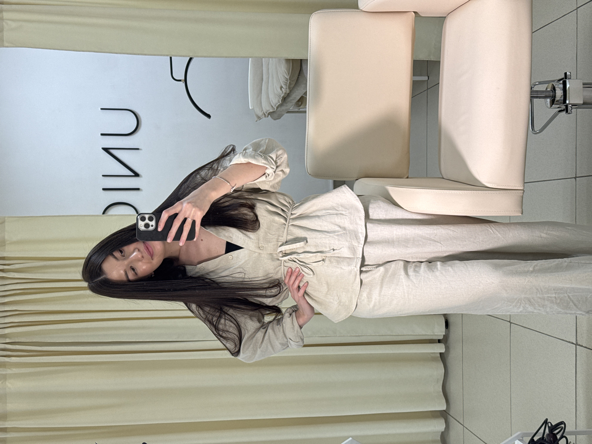 A young woman stands facing a mirror and takes a photo of herself with her smartphone. She is in a room with beige window blinds. A wall with the word "UNIC" is visible in the reflection. Adjacent to the reflection are two high-backed chairs. The floor is tiled. This location is in the city of Chaikovsky, Russia.