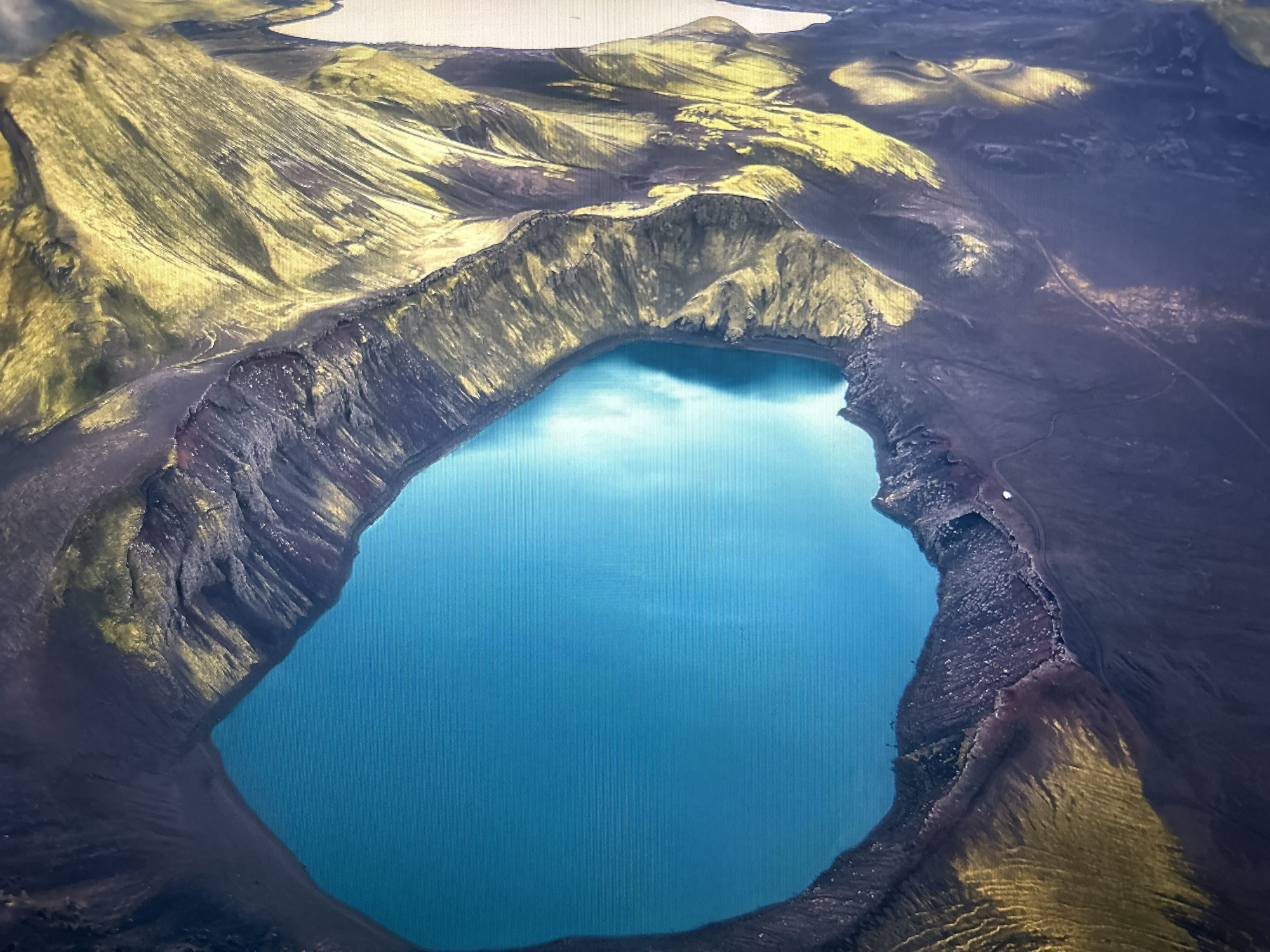 The image is an aerial view of a volcanic crater lake with bright blue water, surrounded by dark, rugged terrain. The crater rim is steep and eroded, with patches of yellowish-green vegetation or mineral deposits. The surrounding landscape consists of rolling hills and plains, also showing evidence of volcanic activity with dark soil and some lighter-colored patches. The overall scene depicts a remote, natural landscape, likely a geological formation in an area with volcanic history. This image is not located in Kuala Lumpur, Malaysia, which is a modern urban city.