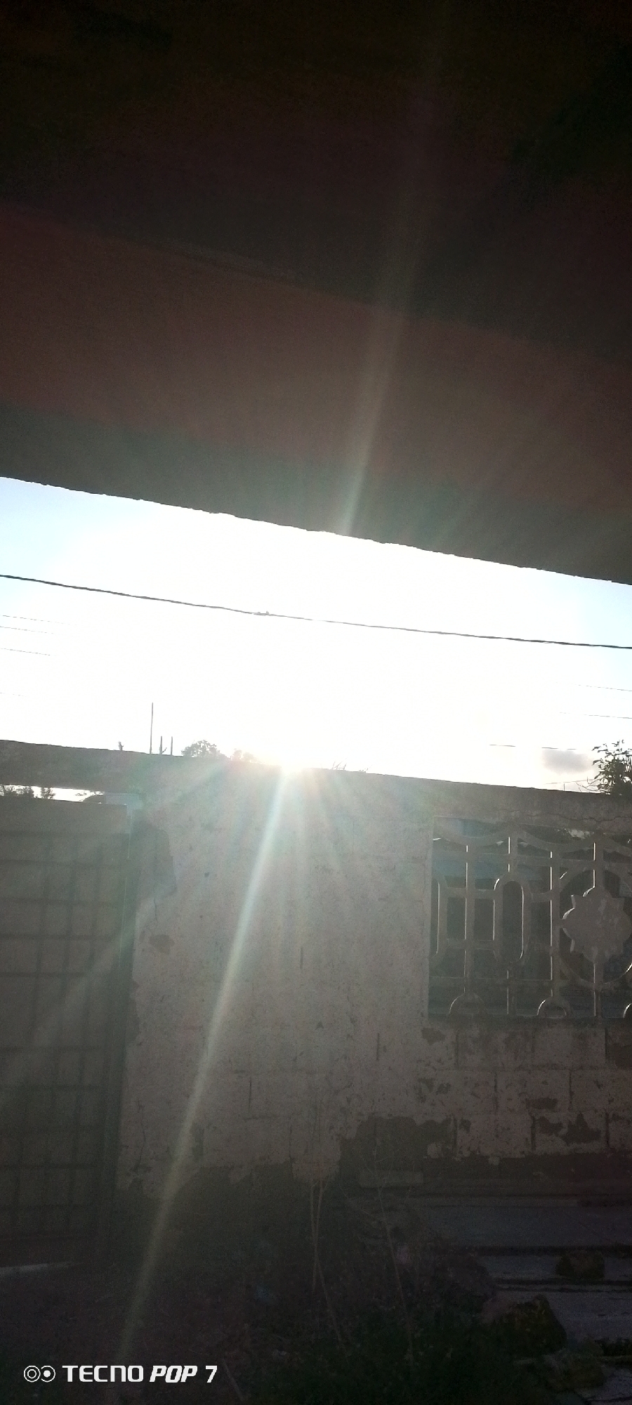 The image depicts an outdoor scene, likely at sunrise or sunset, viewed from a shaded or covered position. A large, dark, horizontal structure, possibly an overhang or roof, dominates the upper part of the frame. Below this, a very bright, clear sky is visible, with thin, dark lines, likely power lines, stretching horizontally across it.
The sun is a prominent feature, positioned centrally in the mid-ground, emitting strong rays and a visible lens flare that spans diagonally across the lower half of the image. Below the bright sky, a light-colored, textured wall, possibly concrete or stucco, with visible wear and patches, extends across the frame. To the left on this wall, a dark, rectangular opening with a grid-like pattern, possibly a gate or window with bars, is present. On the right side of the wall, an elaborately designed metal grill covers a window opening. The bottom of the image shows dark ground, possibly containing sparse vegetation or debris.
No people or specific activities are visible in the static scene. The strong backlighting and lens flare are notable details, indicating direct sunlight from a low angle. The watermark "TECNO POP 7" is visible in the bottom left corner. The scene captures an everyday outdoor environment in Altagracia, Venezuela.