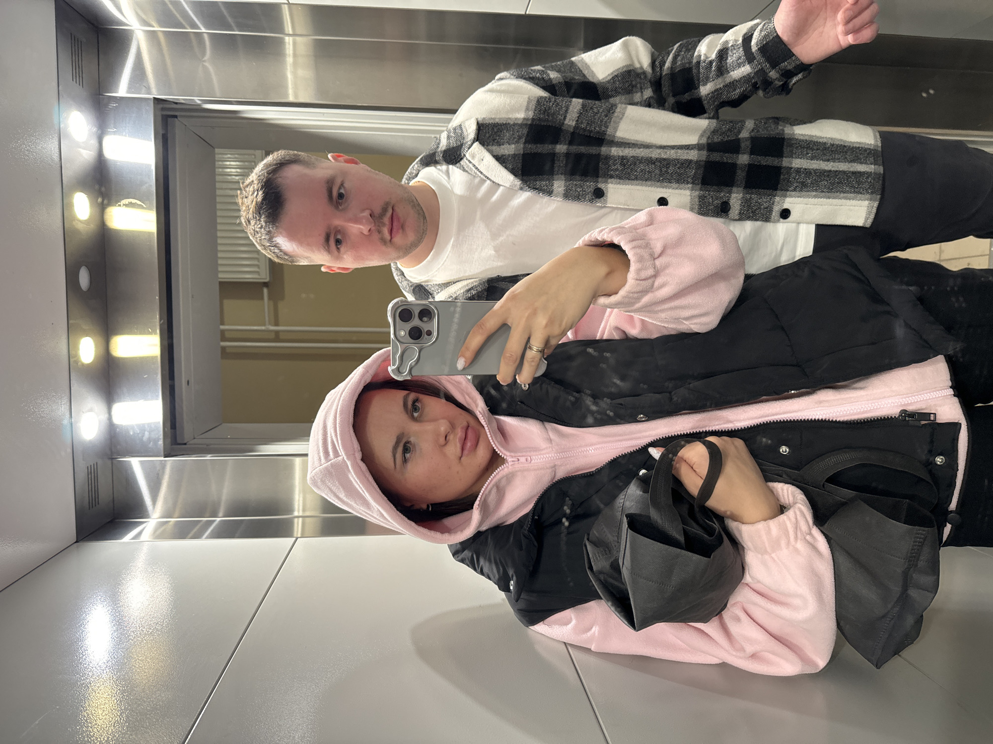 An elevator in Moscow, Russia, contains two individuals: a female and a male. The female, positioned on the left and closer to the camera, is a young adult with light skin and a serious expression. She wears a light pink hooded fleece jacket under a black puffer vest and holds a gray smartphone with a triple-lens camera and a patterned case in her left hand, taking a selfie. A ring is present on her left ring finger. Her right hand grasps a black fabric bag. The male, also a young adult with light skin, stands behind and to the right of the female. He has short light brown hair, a mustache, and a neutral expression, wearing a white t-shirt under a black and white plaid flannel shirt. His right hand is partially visible, held open. The elevator interior features light gray paneled walls and brushed stainless steel door frames and ceiling. Recessed circular lights illuminate the cabin. An open elevator door is visible behind the male, reflecting a lighter-colored room beyond.