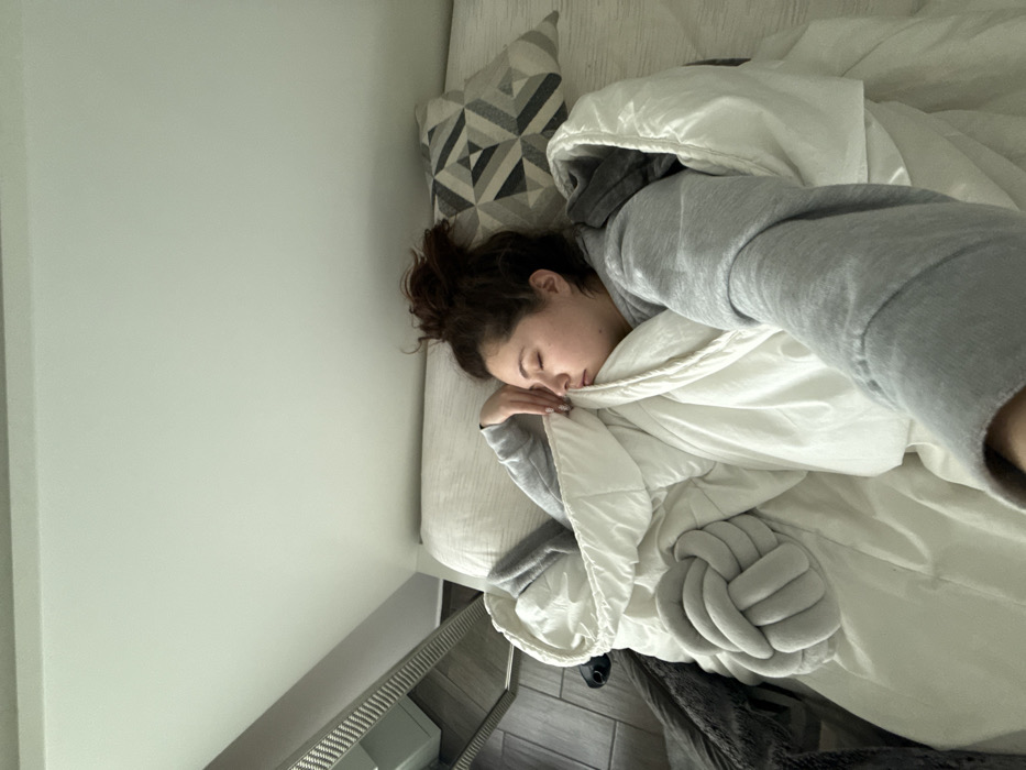 A young adult female is depicted sleeping or resting on her right side in a bed within an indoor bedroom environment, possibly located in Mexico City, Mexico. Her eyes are closed, and her dark brown hair is styled in a messy bun. She wears a light grey hooded sweatshirt and is partially covered by a white comforter. Her left hand rests near her face, while her right arm extends toward the viewer, indicating she is holding the camera. A grey, knotted decorative pillow is visible on the comforter to her left. Behind her head, a pillow with a grey, white, and black geometric pattern is present. The upper portion of the frame shows a plain, light-colored ceiling. To the far left, a section of a grey furniture item with a slatted or ribbed design and a tiled floor are visible. The lighting is bright and even.