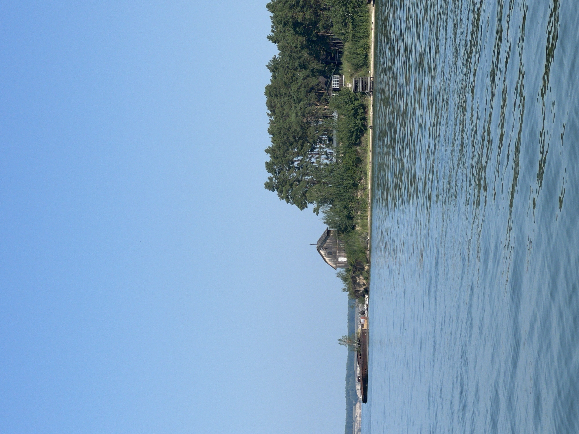 A clear blue sky forms the background of this waterscape scene. The foreground is dominated by rippling blue water. In the middle ground, a narrow strip of land juts out into the water. On this land, a rustic wooden house with a dark roof and a white door stands on a small hill, surrounded by green foliage and tall pine trees. To the left of the house, a derelict, rusty barge is partially submerged in the water, close to the shore. Further along the shoreline, another dark-colored building with a white-paned window is nestled amongst the trees, with a wooden staircase leading down to the water. The shoreline itself is lined with dense green vegetation and sandy patches. The overall impression is one of a quiet, possibly abandoned, lakeside location.
