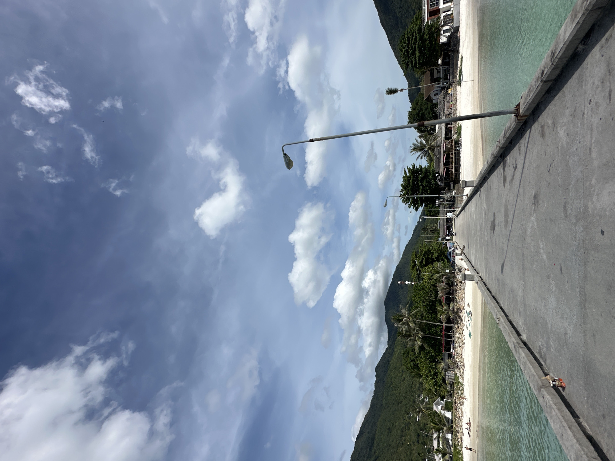 This media file captures a serene coastal scene in Koh Phangan, Thailand. The image is oriented vertically, with the sky dominating the upper left portion and a concrete pier stretching along the right side, leading the viewer's eye towards the horizon.
The pier itself is a prominent object, constructed from rough concrete, with a weathered texture and some rust stains visible near its edge, suggesting it is an established structure. A single lamppost stands along the pier, casting a shadow in the bright daylight. Beyond the pier, a narrow strip of white sand beach is visible, fringed by lush green trees and palm trees, characteristic of a tropical location. In the background, imposing green mountains rise, their slopes covered in dense vegetation.
A few people are scattered on the beach, some appearing to be sunbathing, while others might be walking or enjoying the water. The sea is a clear, inviting turquoise, with gentle ripples indicating calm conditions. The weather appears to be pleasant, with a bright blue sky scattered with fluffy white clouds, suggesting it is daytime, likely late morning or early afternoon given the angle of the shadows. The overall atmosphere conveyed is one of tranquility and relaxation, typical of a tropical beach resort. There is no visible text in the image.