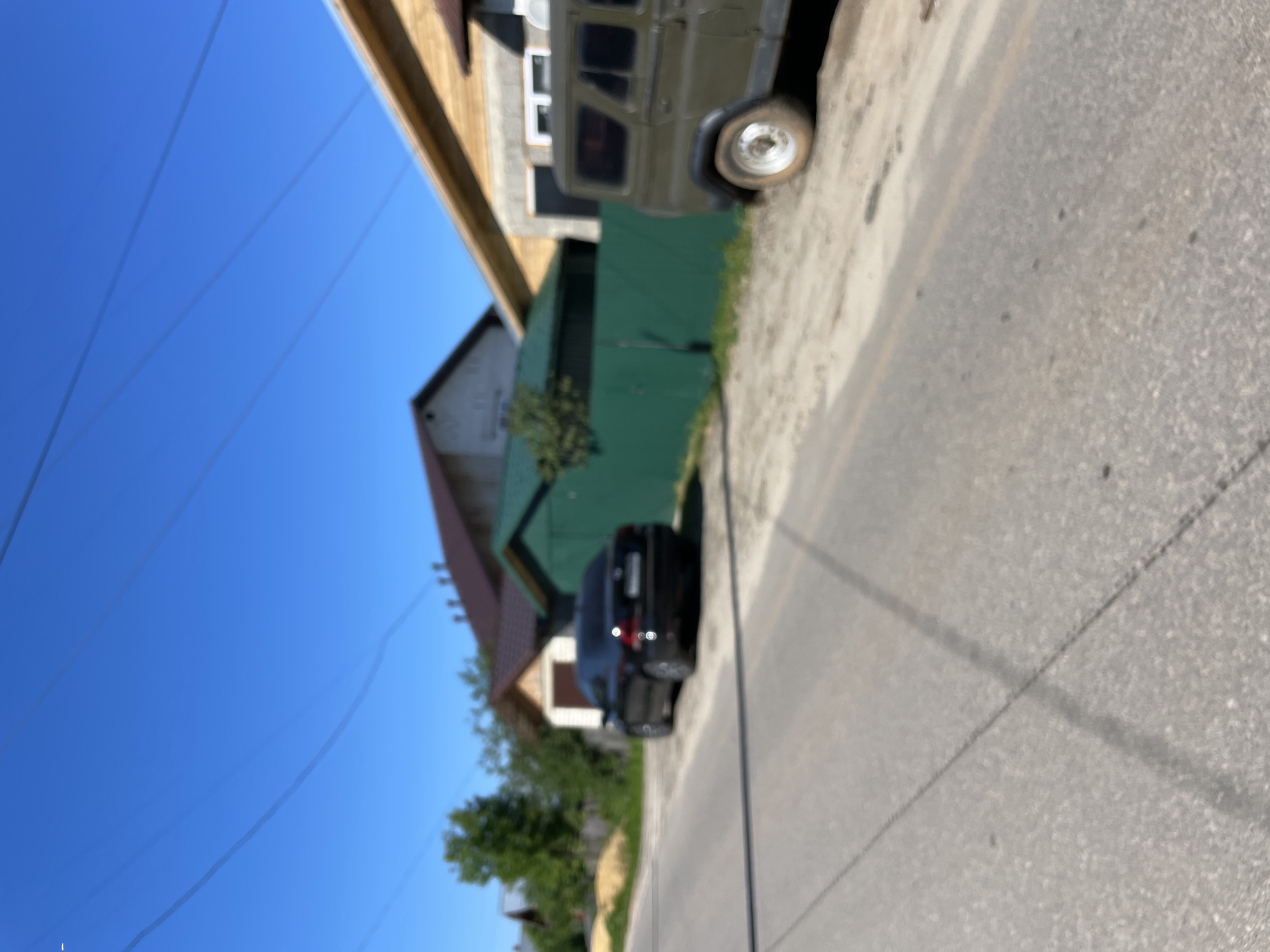 A dark-colored sedan is parked on the side of a paved road in a rural area, with a khaki-colored SUV parked further down the road. In the background, there are several houses with pitched roofs, some with visible windows. A green fence runs along the edge of the properties. The sky is clear and blue, with power lines visible overhead. The scene appears to be located in the settlement of Krasnye Baki, Russia.