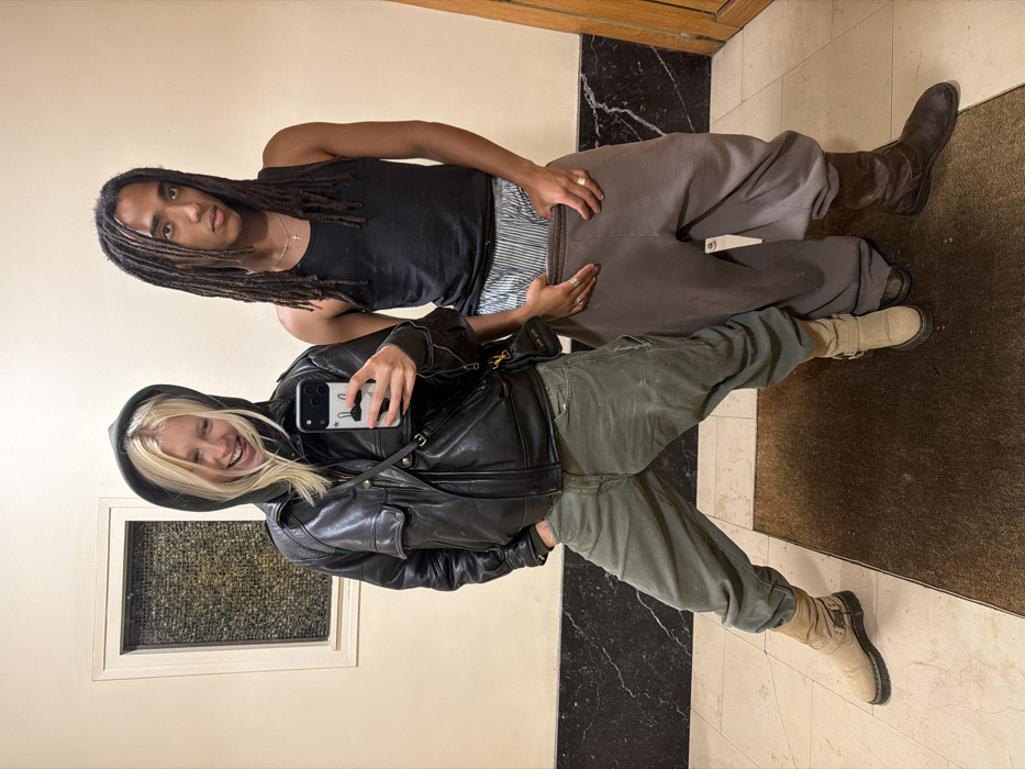 Two people stand in a hallway, taking a selfie. The person in front wears a black leather jacket over a hoodie, green pants, and beige boots, holding a phone with a patterned case. Behind, a person with dreadlocks sports a black tank top, striped boxer shorts, brown sweatpants, and dark brown boots. The hallway features a marble floor pattern, light beige walls, and a window or vent near the bottom left.
