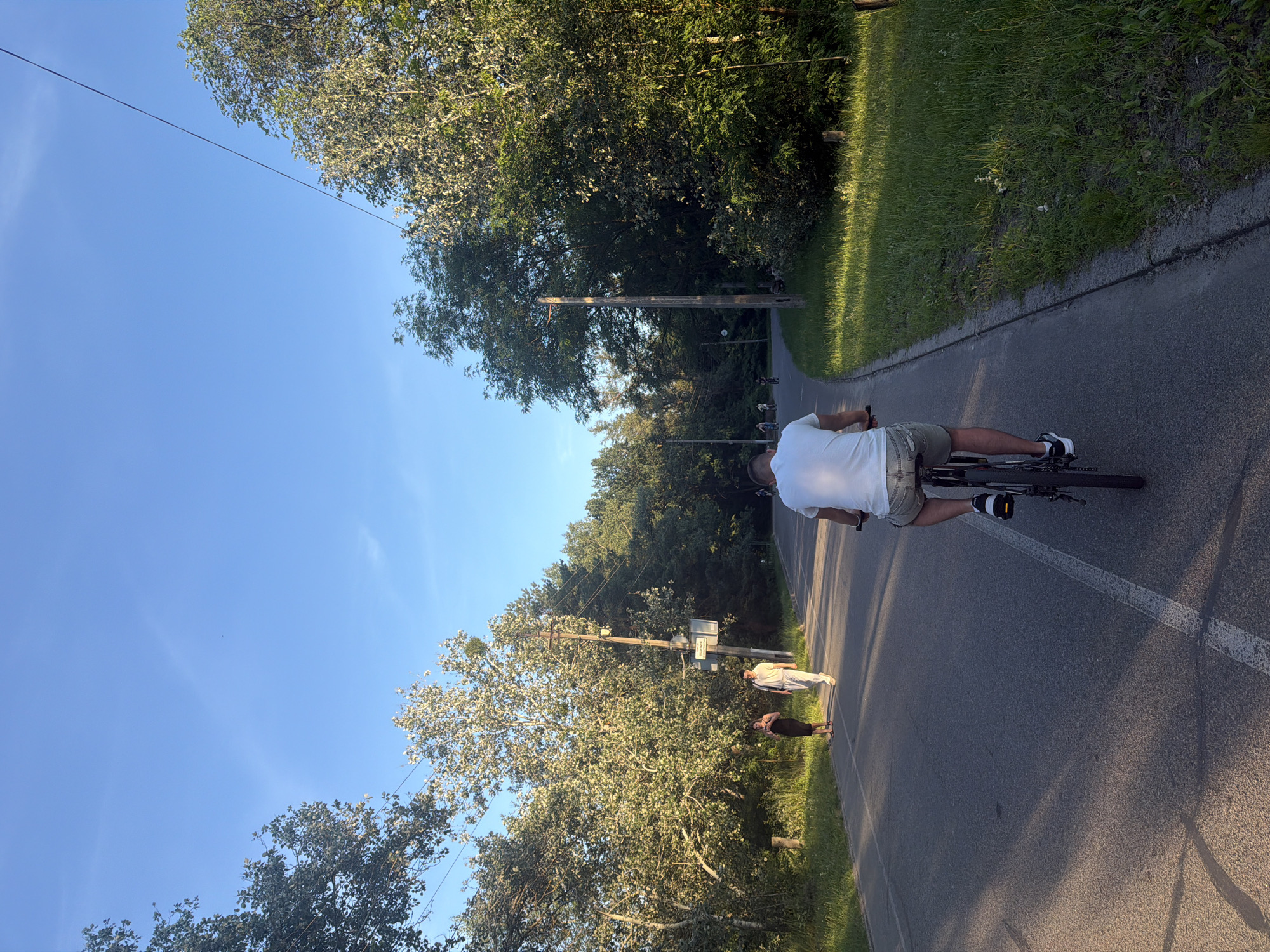 The image depicts a paved path in a park with trees lining both sides and a clear blue sky above. In the foreground, a person is riding a bicycle away from the camera. Further down the path, two individuals are walking, and other people on bicycles are visible in the distance. A utility pole with some signage is situated on the right side of the path. The scene suggests a recreational area within a city park, likely during late afternoon or early evening given the lighting.