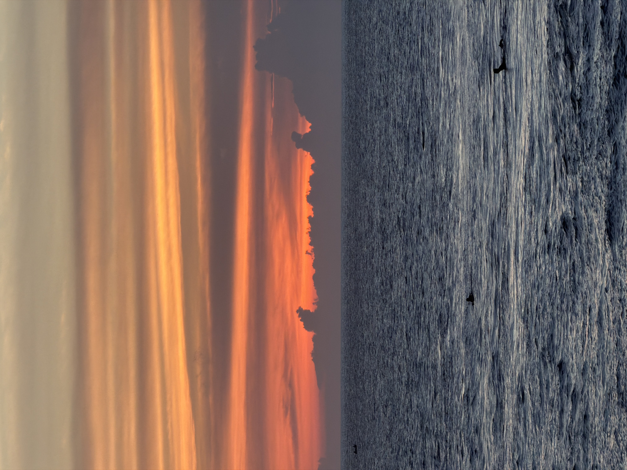 The image shows a vertical split between the sky and the ocean. The sky is a gradient of orange and yellow, with cloud formations visible. The ocean is dark gray, with textured water suggesting ripples or waves. A possible bird is visible in the ocean area. This is likely a beach scene in North Kuta, Indonesia, although no specific landmarks or people are discernible to definitively confirm the location.