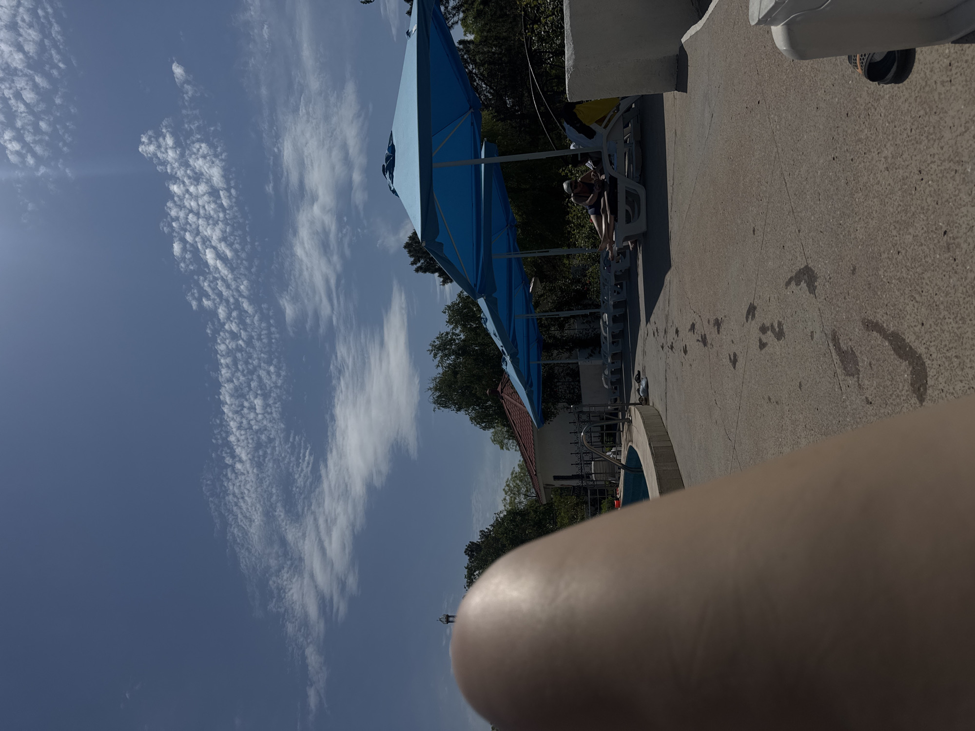 The image shows an outdoor scene, likely at a recreational area with a swimming pool in Yevpatoriya, Ukraine. A row of blue umbrellas shades lounge chairs where individuals are relaxing. A swimming pool is visible nearby, suggesting a leisure setting. The sky is partly cloudy. The image has a close-up of what appears to be an arm or leg in the foreground, obscuring part of the scene. Wet footprints are visible on the concrete surface near the pool. In the background, foliage and a building with a red roof are visible.