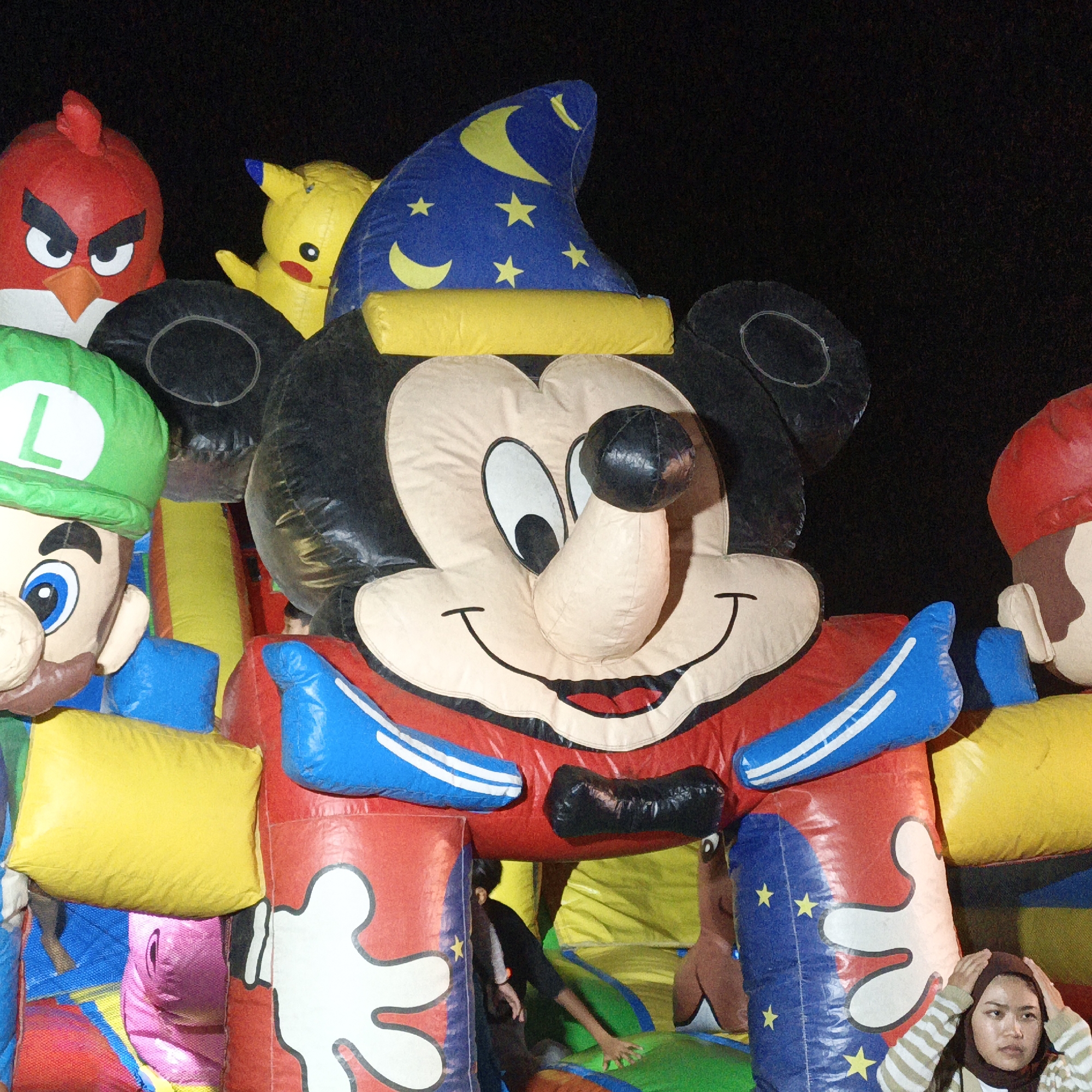 This image depicts a nighttime scene at an inflatable playground in Mangli, Indonesia, likely an event or attraction. The foreground is dominated by a large, inflatable structure featuring characters. Prominently displayed is an oversized inflatable Mickey Mouse wearing a blue wizard's hat adorned with yellow stars and a crescent moon. To the left of Mickey Mouse, a large inflatable Luigi, recognizable by his green hat with an "L" emblem and blue overalls, is visible. Further to the left, a red Angry Bird character with a stern expression is also inflated. To the right of Mickey Mouse, a portion of an inflatable Mario character with a red hat is visible. The inflatable characters are connected and appear to form a large play structure. In the middle ground, a person wearing a dark shirt is partially visible, interacting with the inflatable playground. In the lower right of the image, a young woman wearing a brown hijab and a striped long-sleeved shirt is looking off to the side with her hands on her head, appearing somewhat distressed or contemplative. The background is dark, indicating the photo was taken at night. The overall scene suggests a festive atmosphere, typical of local celebrations or temporary amusement parks.