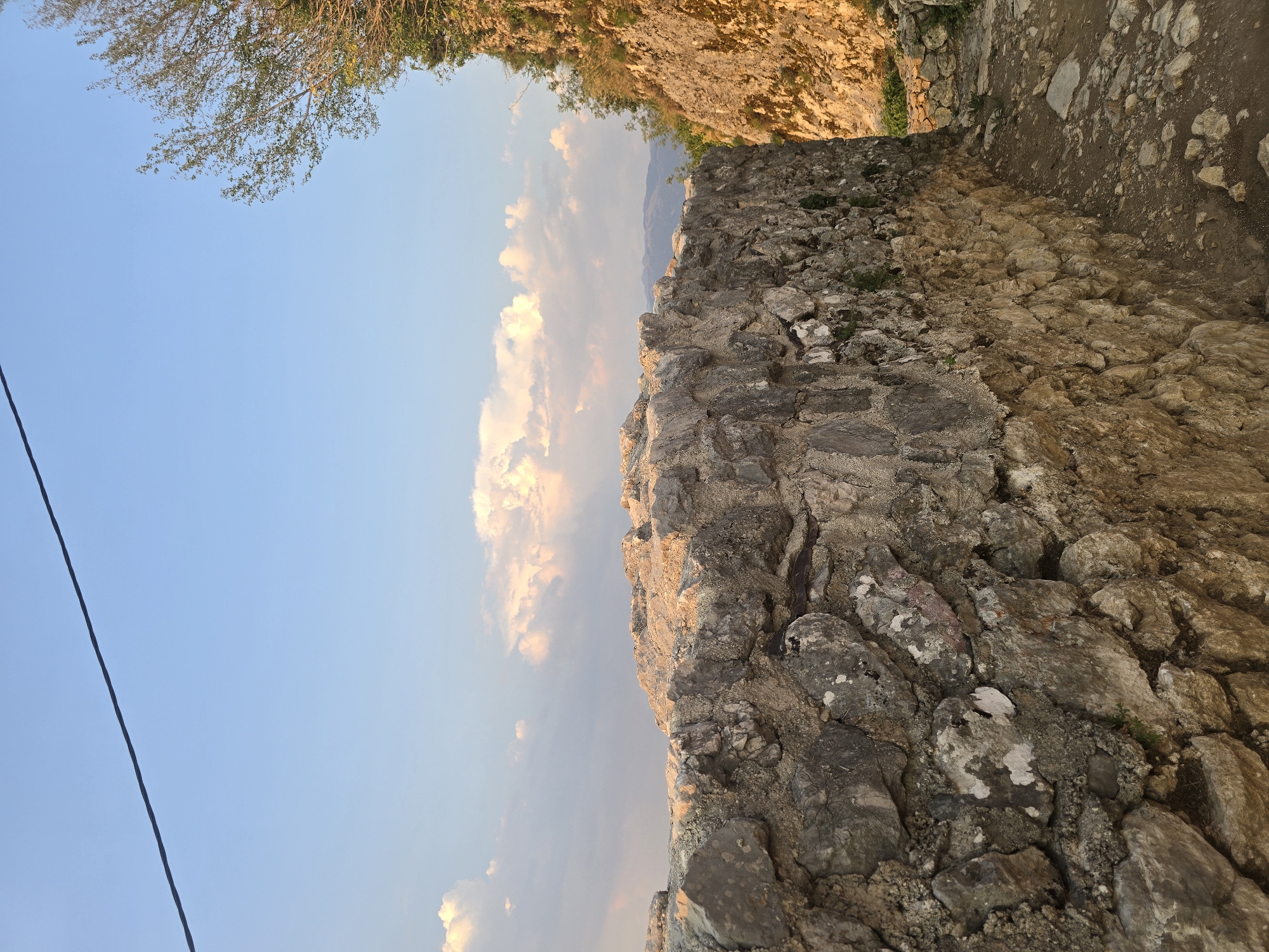 This is a photograph of a stone wall and a path, with a view of the sky and distant hills. The stone wall is rough and appears old, constructed from irregularly shaped gray and brown stones. The path is made of dirt and small stones, winding alongside the wall. To the right of the wall, a steep, rocky slope covered in greenery is visible, bathed in warm, late afternoon sunlight. Above, the sky is a clear blue, with scattered, fluffy white clouds tinged with the orange glow of sunset. A thin black wire stretches diagonally across the upper portion of the frame. The scene appears to be outdoors, likely in a mountainous or hilly region. The location is identified as Gornja Vrbica, Montenegro. There are no people or animals visible in the image.