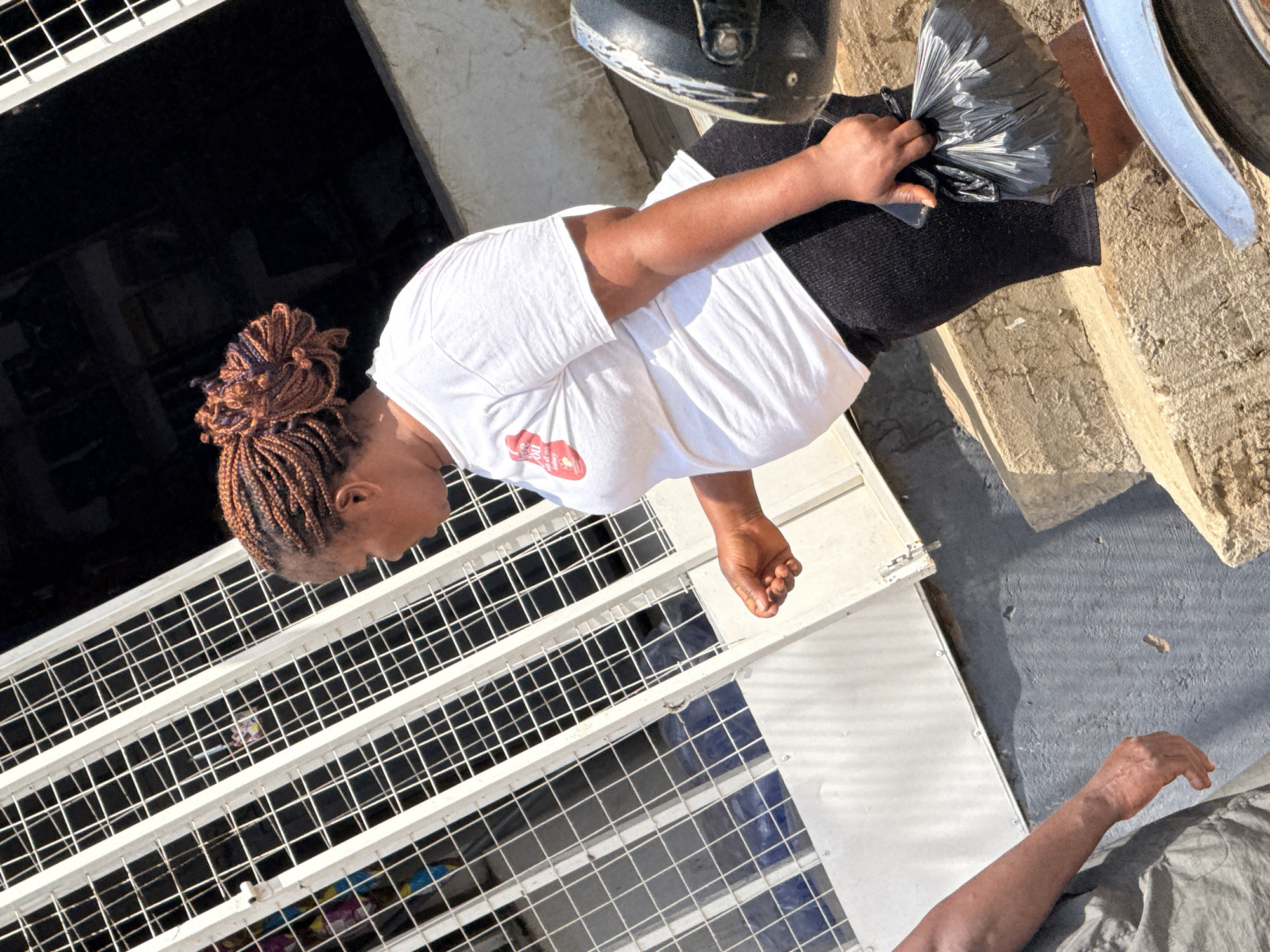 An outdoor, mid-shot, eye-level, vertical, full-color photograph depicts a woman standing in front of a white metal gate. The woman, who is of African descent, has braided brown hair styled in a bun and is wearing a white t-shirt and black shorts. She is holding a black plastic bag in her right hand. To her left, a person wearing a gray outfit is partially visible, with their arm extending into the frame. The background features a textured wall and concrete steps. The scene appears to be in an urban setting, possibly a market or street in Abuja, Nigeria, given the architectural style of the gate and the general environment. The lighting suggests it is daytime, with sunlight casting shadows.