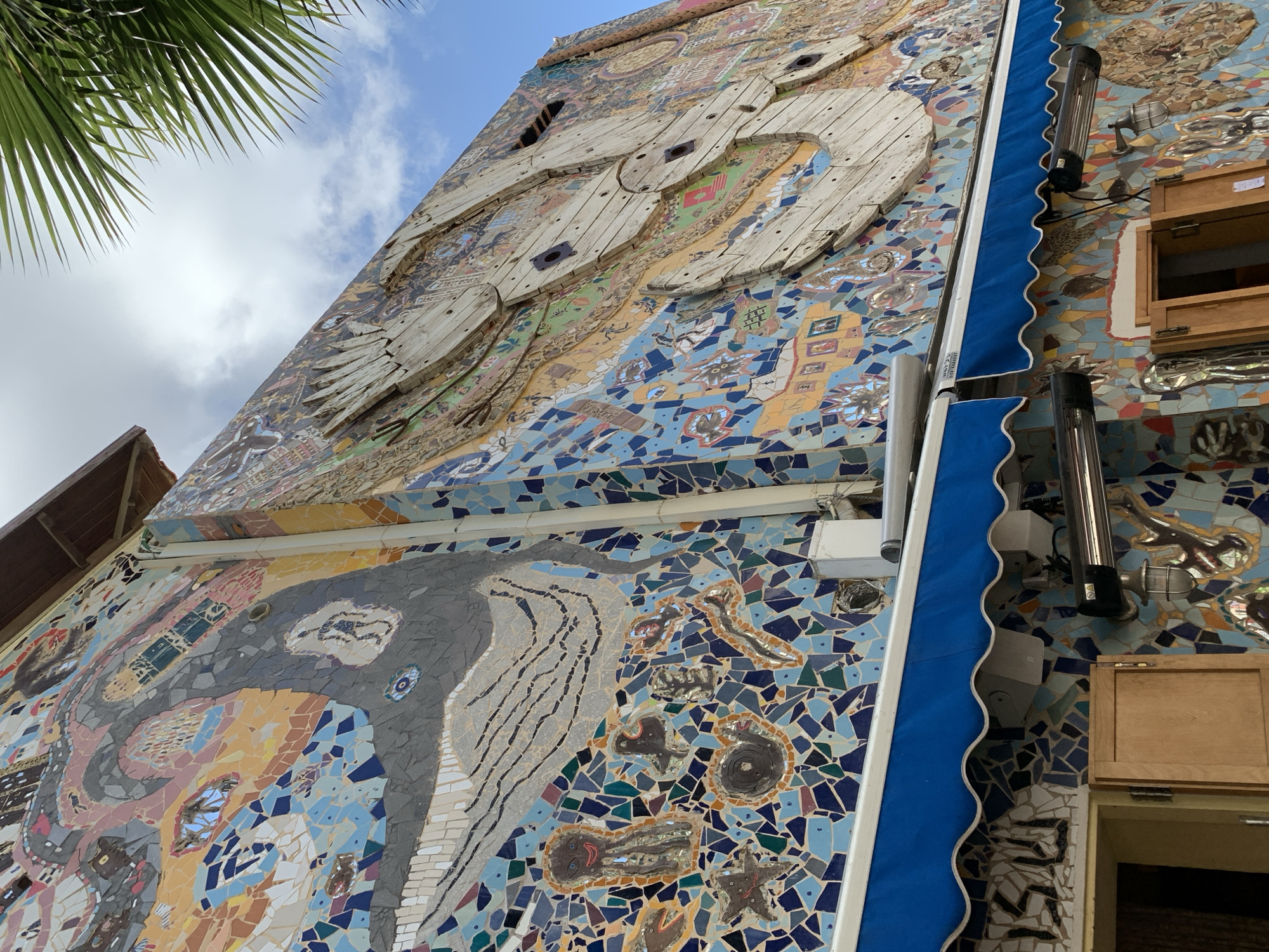 Here is a detailed description of the image:
The image depicts a building facade in Tel-Aviv, Israel, covered in mosaic art. The artwork features marine life, including a whale and a bird constructed from reclaimed wooden spools. The mosaic incorporates a variety of materials, with blue, turquoise, and neutral tones dominating the color palette.
Attached to the building are practical elements, such as a blue awning, an external heating unit, and wooden cabinets. A palm tree is visible in the upper left corner of the frame, suggesting an outdoor, possibly coastal, environment. The sky is visible in the background, showing partial cloud cover.