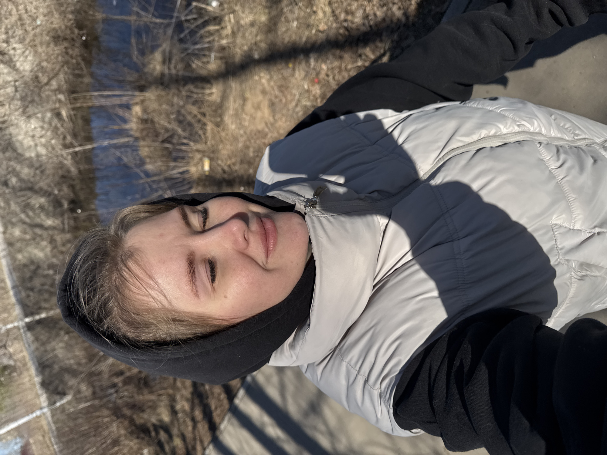 A young woman is lying down outdoors. She is wearing a light gray puffer jacket over a black hooded sweatshirt. The background shows a body of water with dry vegetation along the bank. The location appears to be the city of Ферма, Russia. The lighting is bright, suggesting it is daytime. The woman's eyes are closed, and she has a relaxed expression. The scene is static.