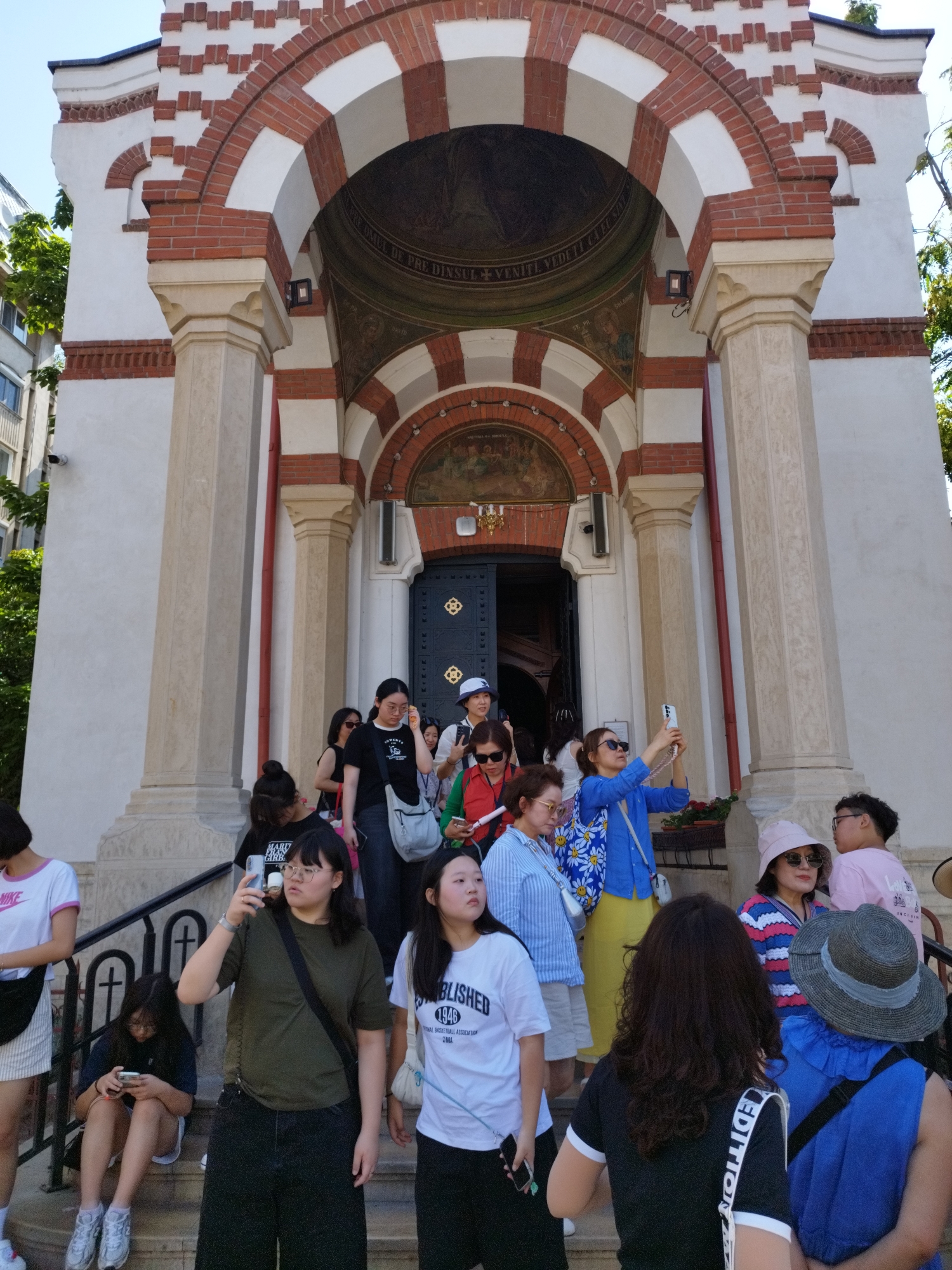 A group of tourists is gathered on the steps of a building that appears to be a church or a historical monument. The architecture features red and white brick arches and columns. The tourists are of various ages and ethnicities, and many are holding cell phones, suggesting they are taking pictures or videos.
Several individuals are prominently featured:
- A young woman in a white t-shirt with "NIKE" logo and a white skirt is on the far left, looking away from the camera.
- A young woman in a dark green t-shirt and dark pants is in the foreground, holding a cell phone up to take a picture.
- Another young woman in a white t-shirt with "ESTABLISHED 1946" text is to the right of the previous woman, looking off to the side.
- A woman in a blue shirt and yellow pants is in the middle ground, holding a cell phone and seemingly taking a selfie.
- A woman in a striped pink and blue sweater and a pink hat is on the right side, looking to the side.
- A person wearing a large, grey brimmed hat and a blue top is in the foreground on the right.
The environment suggests a warm, sunny day, with trees visible in the background. The overall scene depicts a moment of tourism and cultural exploration.
This building is the **Stavropoleos Monastery Church** in Bucharest, Romania. It is known for its distinctive architecture, incorporating elements of Brancovenesc style and Byzantine influences. The small church, founded in 1724, is a popular landmark in the historic center of Bucharest.