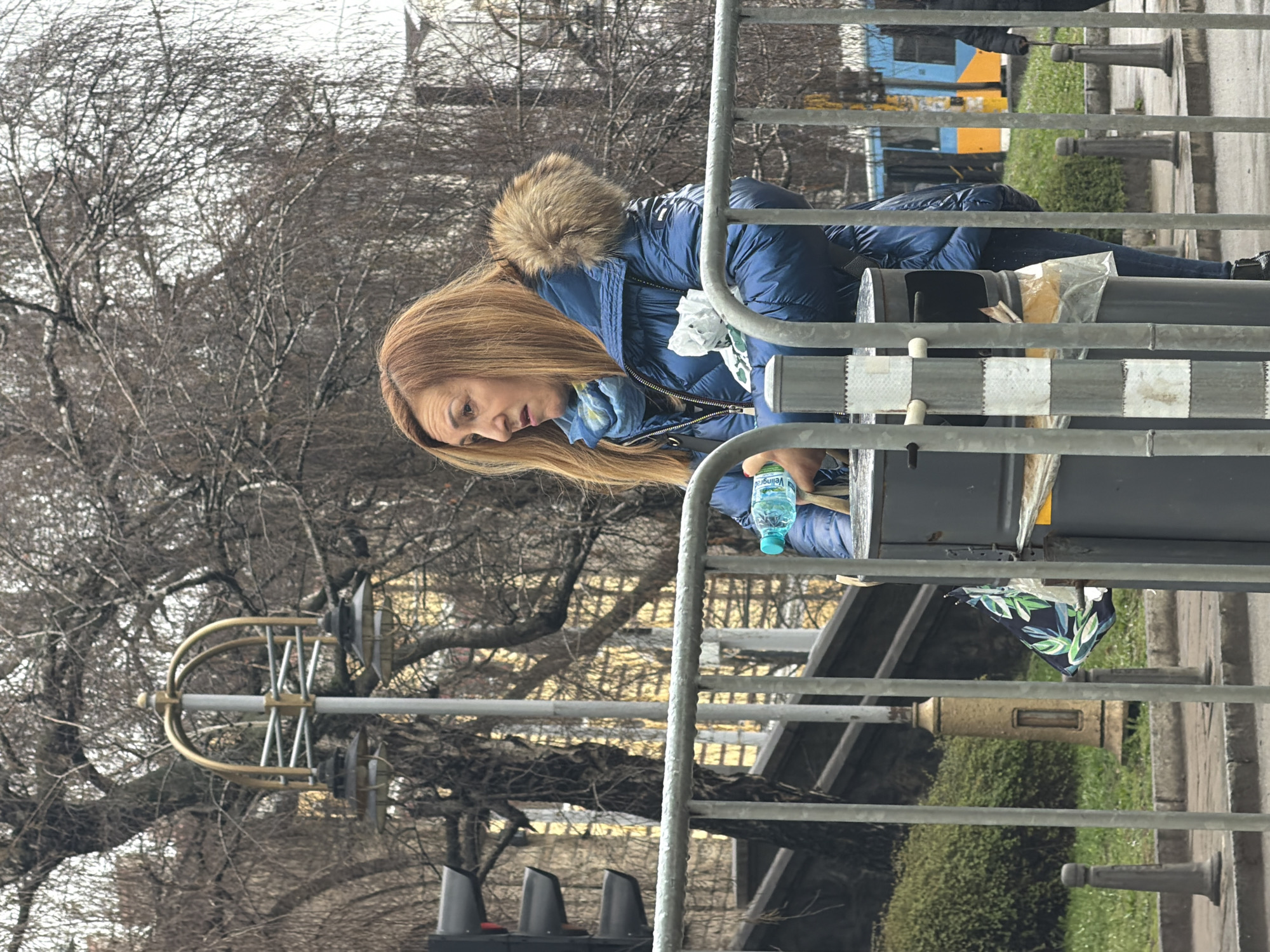 The image captures a street scene in central Sofia, Bulgaria, under an overcast sky with bare deciduous trees, indicative of late autumn, winter, or early spring. In the mid-ground, a woman with long, reddish-brown hair is positioned slightly to the right of a dark grey public trash bin. She wears a shiny blue puffer jacket with a light brown fur-lined hood and a blue patterned scarf. In her right hand, she holds a clear plastic water bottle with a blue cap and a green label, displaying "Veilingrad." Her left arm appears to cradle a crumpled white item and a black and green patterned umbrella or bag. Her mouth is slightly open, and her gaze is directed intently towards her left, off-camera, suggesting a reaction or conversation.
A grey metal crowd barrier occupies the immediate foreground, partially obscuring the woman and the trash bin. To the far left, a black traffic light pole supports three horizontal signal lights. Directly behind the woman, a distinctive streetlight features a grey pole topped with an ornate, gold-colored arched fixture holding two downward-facing lamps. Further in the background, a portion of a yellow-orange and blue tram or trolleybus is visible on the right, traveling leftward. The environment includes a grey sidewalk with pedestrian bollards, green ground cover, and a set of concrete steps or a raised pathway.