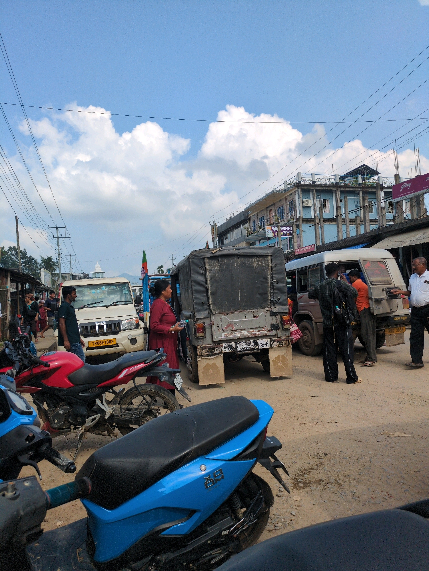 In Kanchanpur, India, a daytime scene unfolds on a dusty road with several vehicles and people. A white SUV with license plate TR05E1883 is parked, and a man in a dark green shirt stands beside it. To the right, a covered military-style truck is present, with a woman in a maroon outfit holding a bottle of water near its rear. A light-colored van, possibly a passenger vehicle, is positioned to the right of the military truck.
Further into the scene, a red motorcycle is parked prominently in the foreground, and a blue scooter is partially visible in the lower left corner. Behind the light-colored van, a man in an orange shirt is interacting with someone inside the van. A man in a black and green checkered shirt, wearing a backpack, is also near the van. A man in a white shirt and dark pants stands to the far right, gesturing.
The background features a multi-story building with blue accents and balconies, and a smaller structure with a sloped roof. Overhead, power lines crisscross a partly cloudy blue sky. The ground is a mix of dirt and gravel, indicating an unpaved or poorly maintained road surface. Several other figures are visible in the distance, suggesting a populated area.