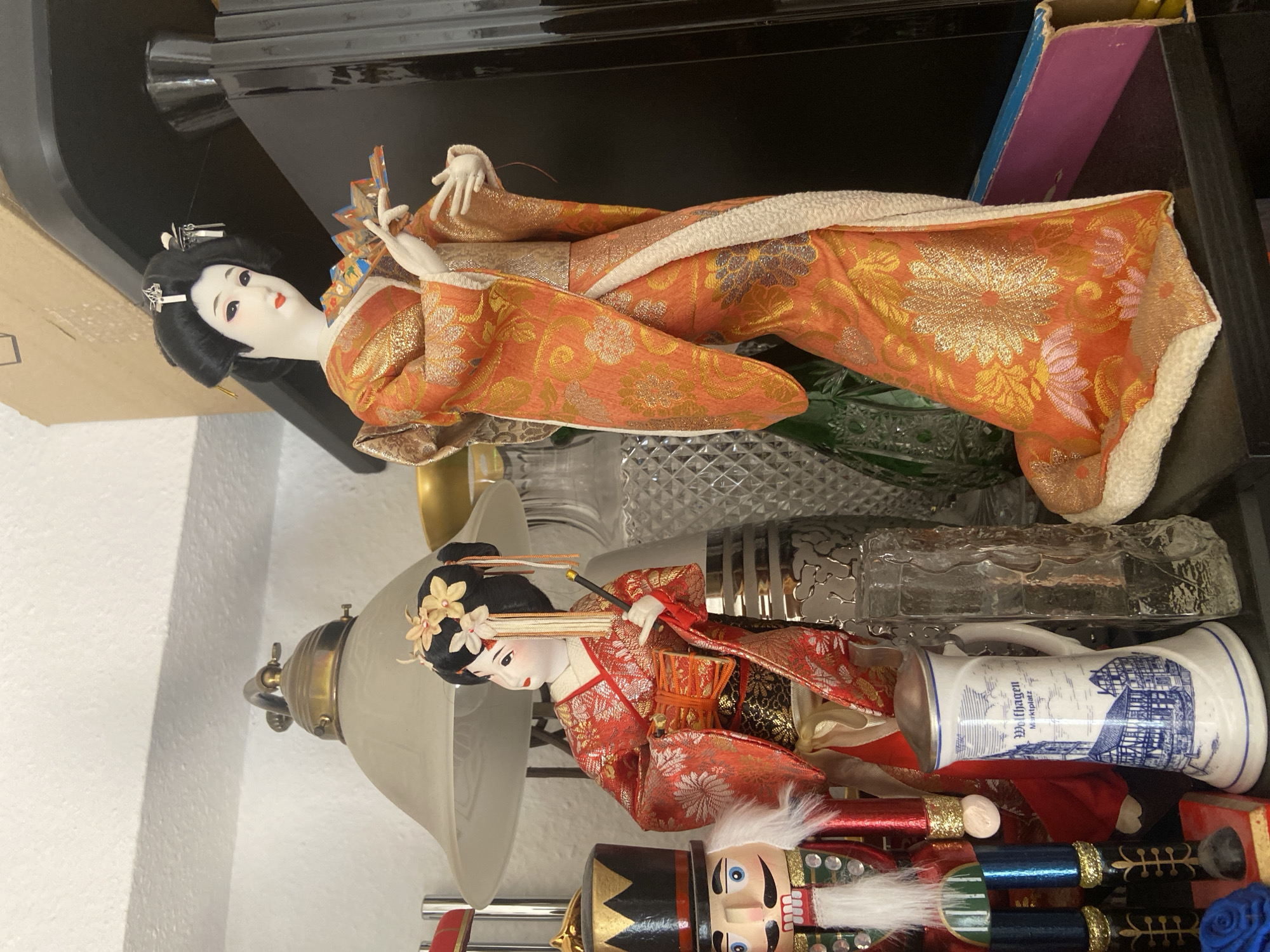 The image displays a shelf with various decorative items. On the right side of the shelf is a large Japanese geisha doll in an orange kimono adorned with gold floral patterns. She is holding a folded fan in her right hand. To the left of the geisha doll is a smaller Japanese doll, also dressed in a red and gold kimono, holding a drum. In front of these dolls, on the left side of the shelf, stands a nutcracker figurine dressed in a red and green uniform with a black hat. Next to the nutcracker is a ceramic beer stein with a blue and white illustration of buildings and the text "Wolfhagen Marktplatz". This illustration depicts buildings in the town of Wolfhagen, which is located within the city of Kassel, Germany. Behind the dolls and the beer stein are various glass objects, including a textured green bottle and a clear decorative bottle. A vintage lamp with a frosted glass shade is visible in the upper left portion of the image. The background consists of a white textured wall and a black shelf.
