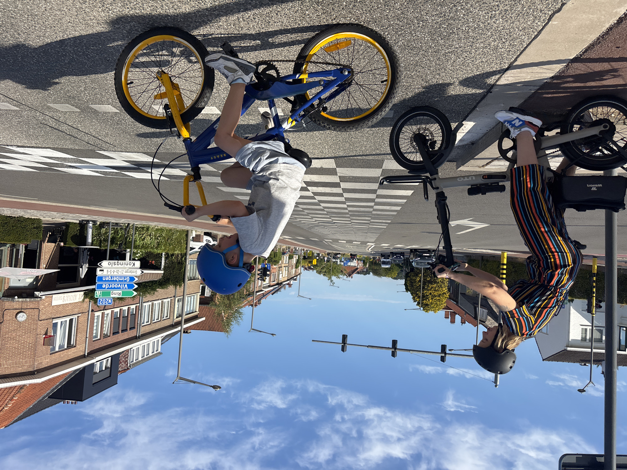 This image shows two individuals on bicycles, facing away from the camera, in a suburban street setting. The person on the left is a child on a blue and yellow BMX-style bike with yellow rims and blue frame. They are wearing a blue helmet and a light gray t-shirt. The child is in motion, with their legs pedaling.

To the right, an adult is on a lighter colored bicycle, possibly an e-bike, with a black basket attached to the rear. This person is wearing a black helmet and a brightly striped outfit with orange, yellow, black, and blue stripes. Their left leg is raised, resting on the pedal.

The scene is a paved road with a pedestrian crossing marked by white and black checkered patterns. In the background, there are brick houses with tiled roofs, trees, and traffic lights. Street signs are visible, including one with "N202" and "Vilvoorde" written on it, indicating the location as Vilvoorde, Belgium. The sky is blue with some white clouds, suggesting it is daytime, likely sunny and warm. The overall mood appears to be one of leisure and outdoor activity.