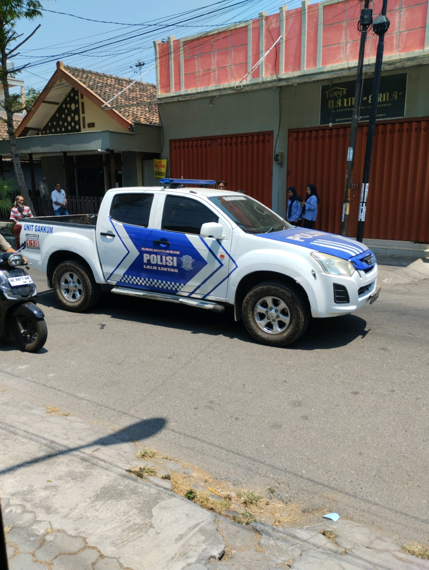 A white Isuzu D-Max pickup truck, marked with "POLISI" and "LAUL LINTAS" in blue lettering, is parked on the right side of a paved road in front of buildings. The truck has blue and white graphics on its sides and a blue light bar on the roof. Two women in blue uniforms are standing on the sidewalk near the truck. A black motorcycle is parked on the left side of the road in front of the truck. A man with a striped shirt and a man in a white shirt and blue jeans are standing on the sidewalk behind the motorcycle. The buildings have red corrugated metal doors and a sign that reads "GRIYA BATIN SERNA." The scene appears to be in Ponorogo, Indonesia.