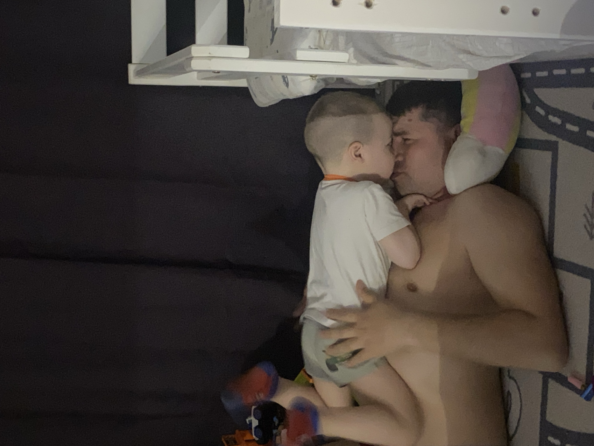 A father and son are lying together in a room. The father is shirtless and lies on a bed covered with a gray patterned sheet. A child wearing a white shirt and gray bottoms is on top of the father with his face near the father's face. Toys are on the floor. White furniture is visible above them. The background is black. The location is unknown and no connection to the city of Sochi, Russia is visible.