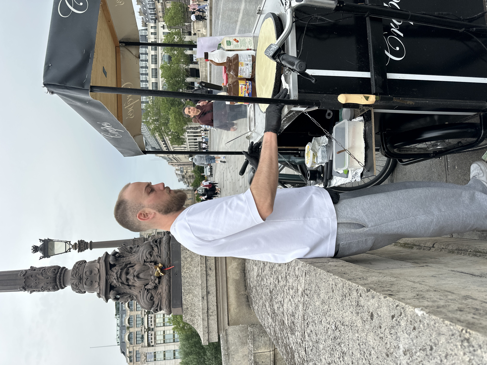 The image depicts a man standing at a crepe stand in Paris, France. The man is fair-skinned with short hair and a beard. He wears a white t-shirt and gray sweatpants and black gloves. He is positioned at a black crepe cart with a flat, round griddle, appearing to be in the process of making a crepe. The cart has the word "Crêpes" written in white cursive letters on its side. Various ingredients and supplies such as Nutella, milk, and butter are visible on the cart. In the background are Parisian buildings, trees, and pedestrians. The scene is captured on a gray, overcast day. The photograph is taken at eye level. A decorative stone lamp post is in the foreground.