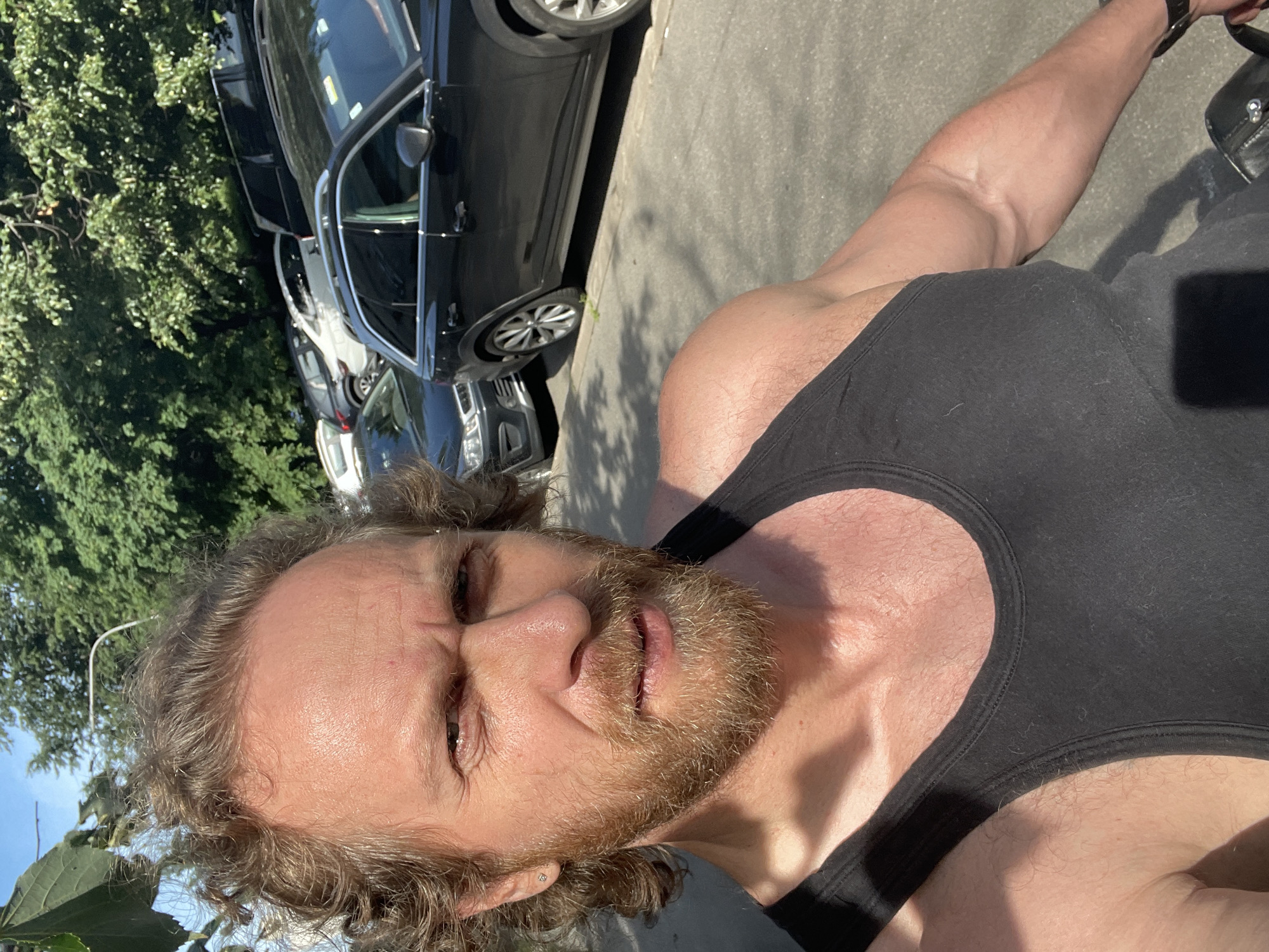 A man with curly brown hair and a beard, wearing a black tank top, takes a selfie outdoors. He has a diamond stud earring in his left ear. In the background, several cars are parked on the side of a street, with trees and a blue sky visible above. The scene appears to be a sunny day in Saint Petersburg, Russia.